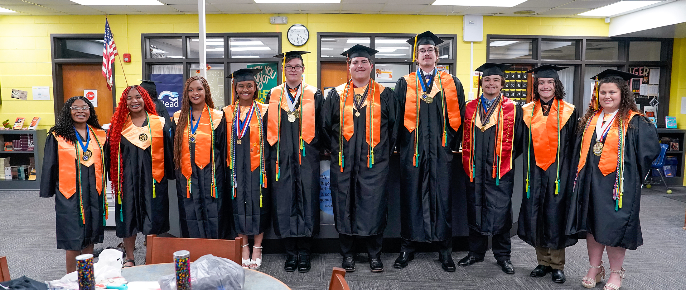 A group of graduates in black gowns and orange sashes stand in a library with bookshelves in the background.