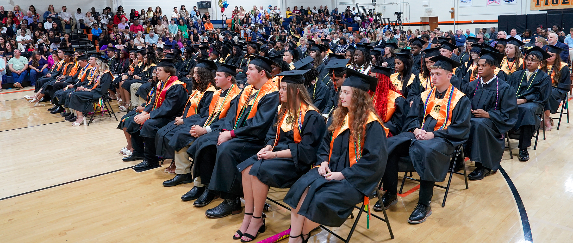 Graduates in black gowns and caps with orange sashes sit in the foreground, while a large audience fills the bleachers in the background.