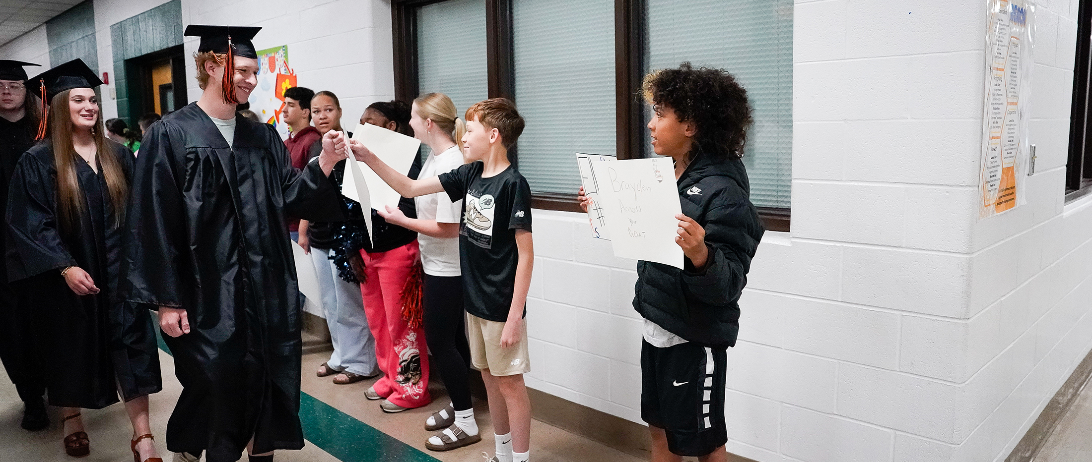 A group of students in graduation gowns and casual attire stand in a hallway, with one student in a graduation gown handing a paper to another student.