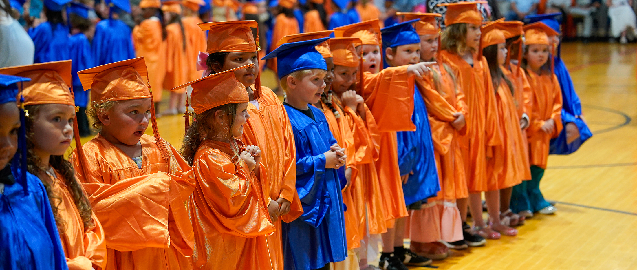 A group of young children in orange and blue graduation gowns and caps stand in a line, with a blurred background of more graduates.