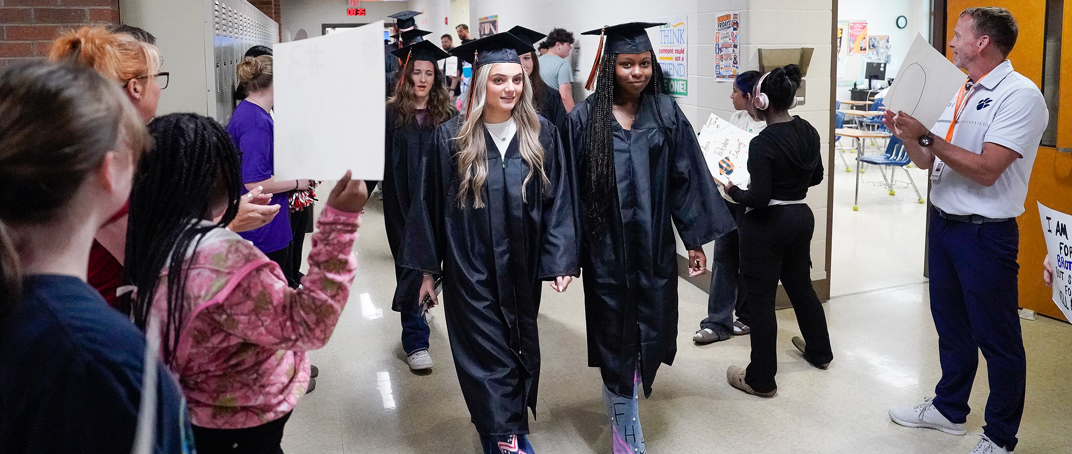 Two graduating students in caps and gowns walk down a hallway lined with cheering onlookers holding signs.