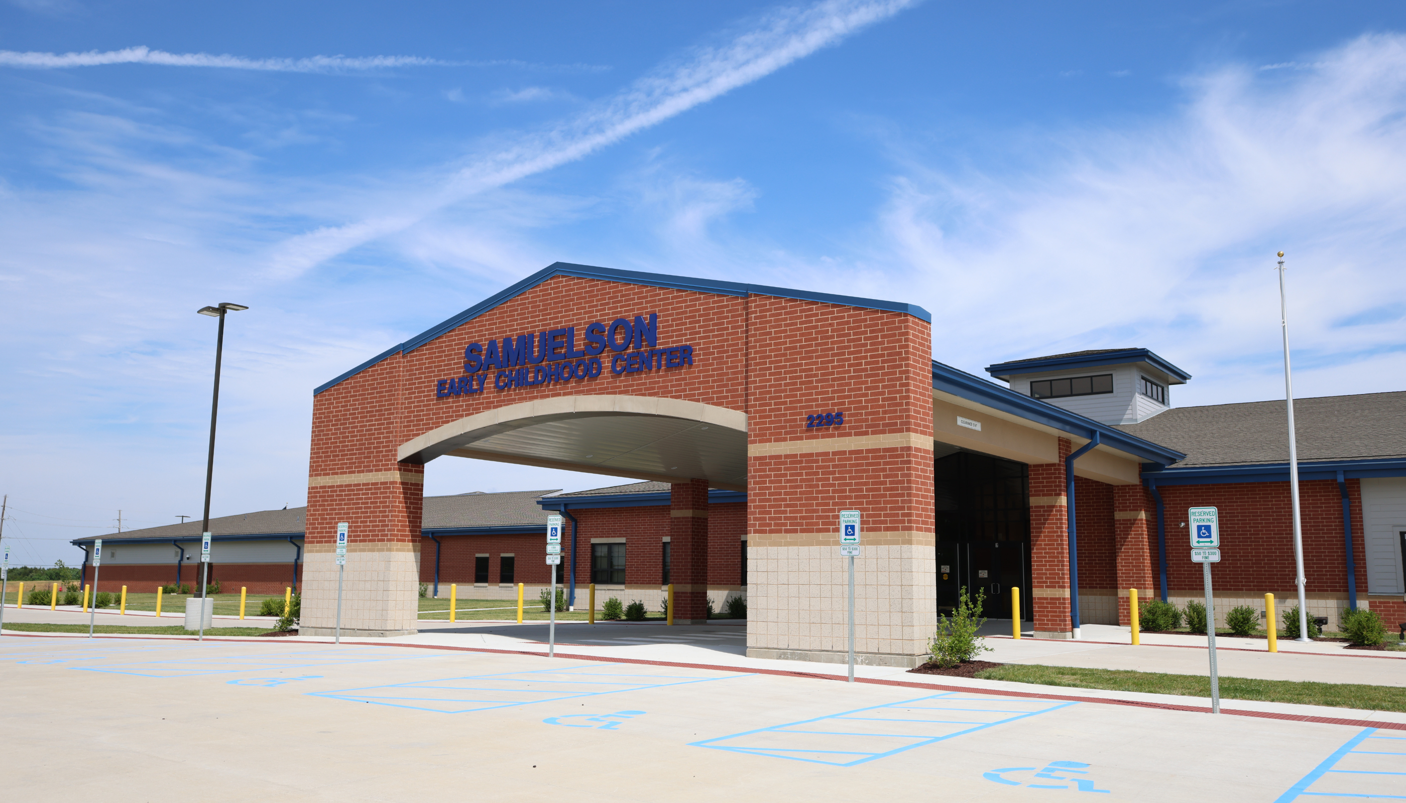 A bright blue sky with wispy clouds stretches over the red brick entrance of the Samuelson Early Childhood Center, with a paved parking lot in the foreground.