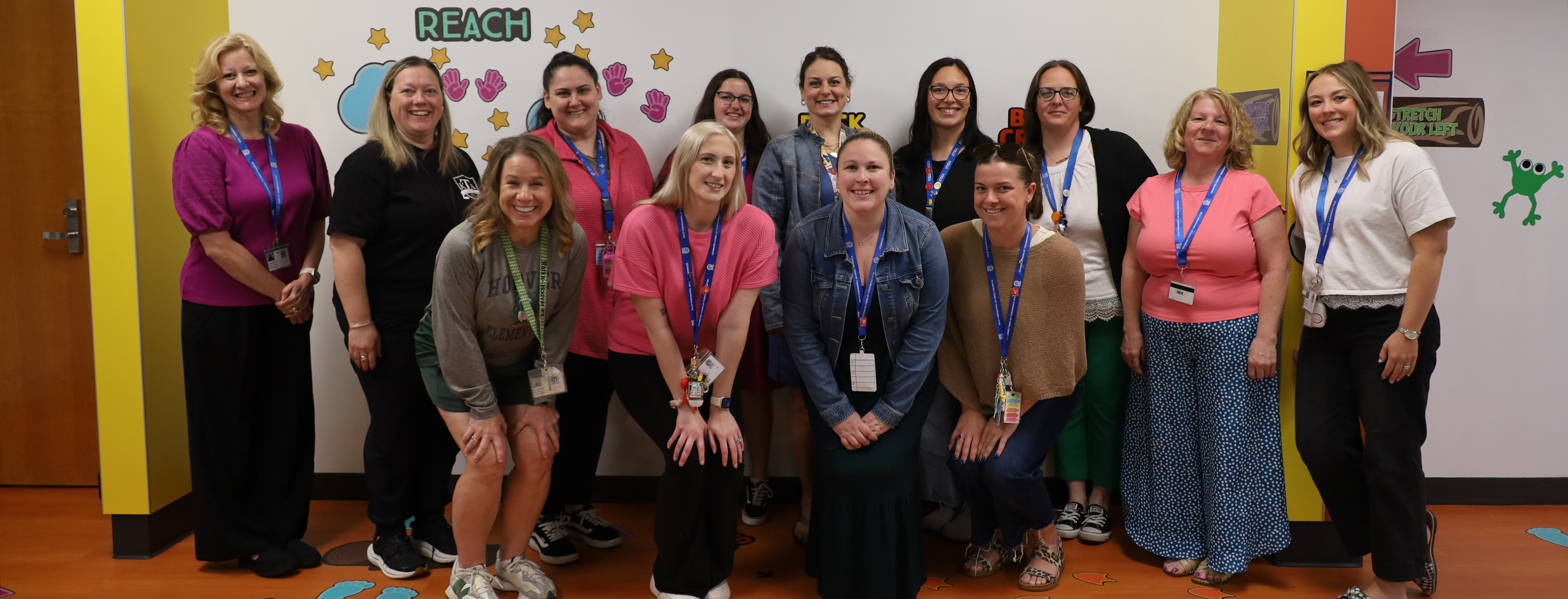 A group of women stand in front of a colorful wall with the word "REACH" and stars, while a few women are in the foreground.