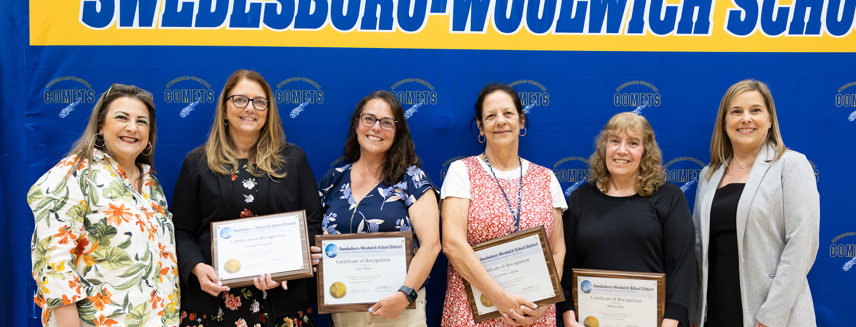 Six women stand in front of a blue banner with yellow lettering that reads "SWEDESBORO-WOOLWICH SCHOOLS," holding certificates.