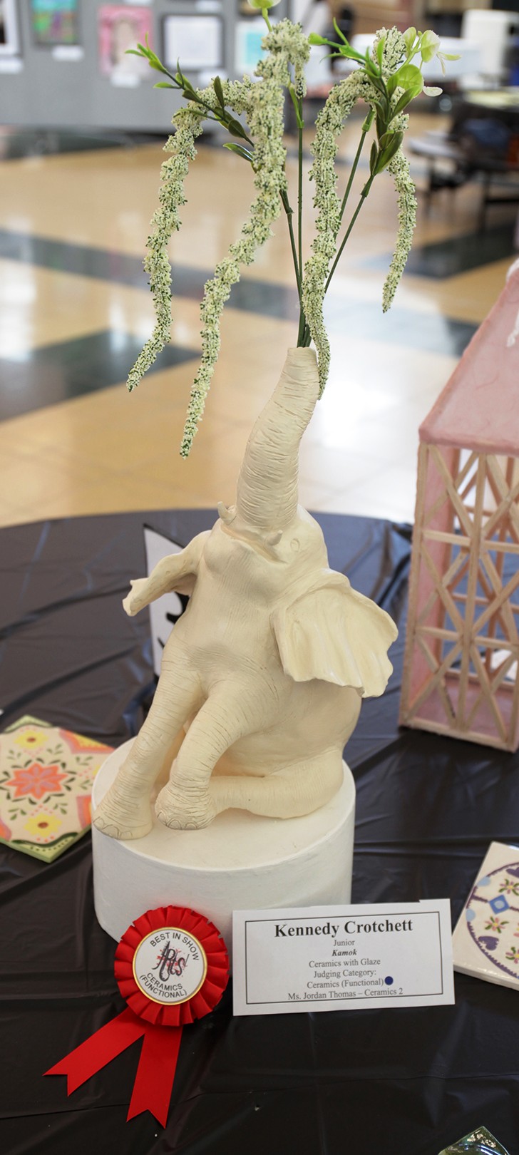 A ceramic elephant sculpture with a red ribbon and award label in the foreground sits on a black tablecloth, with a pink geometric structure and tiled artwork in the blurred background.