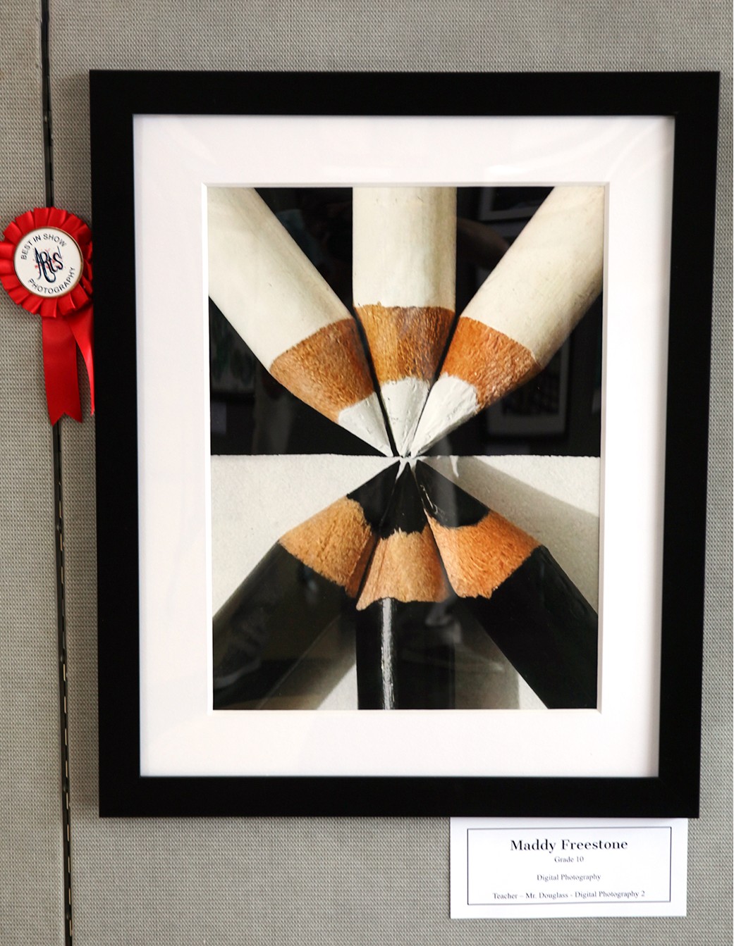 A framed photograph of sharpened pencils arranged in a starburst pattern against a black and white checkered background is displayed with a red ribbon and a label.