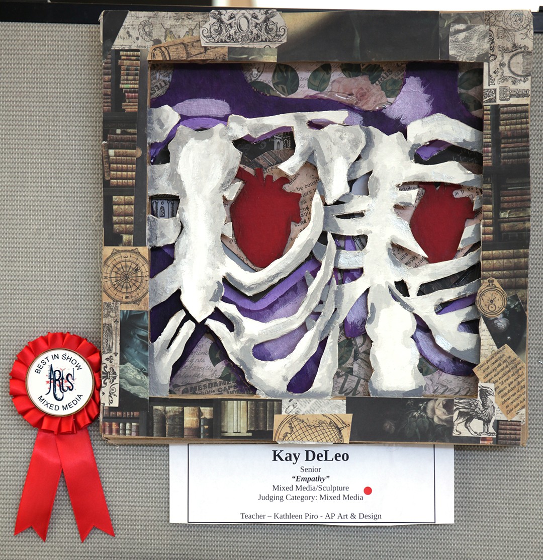 A mixed-media sculpture of a ribcage with red hearts is displayed in a shadow box adorned with book-themed paper, with a "Best in Show" ribbon in the foreground.