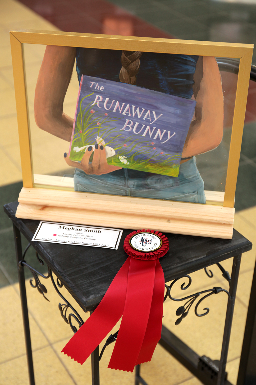 A framed illustration of "The Runaway Bunny" is displayed on a table with a red ribbon and award, set against a blurred background of a building interior.