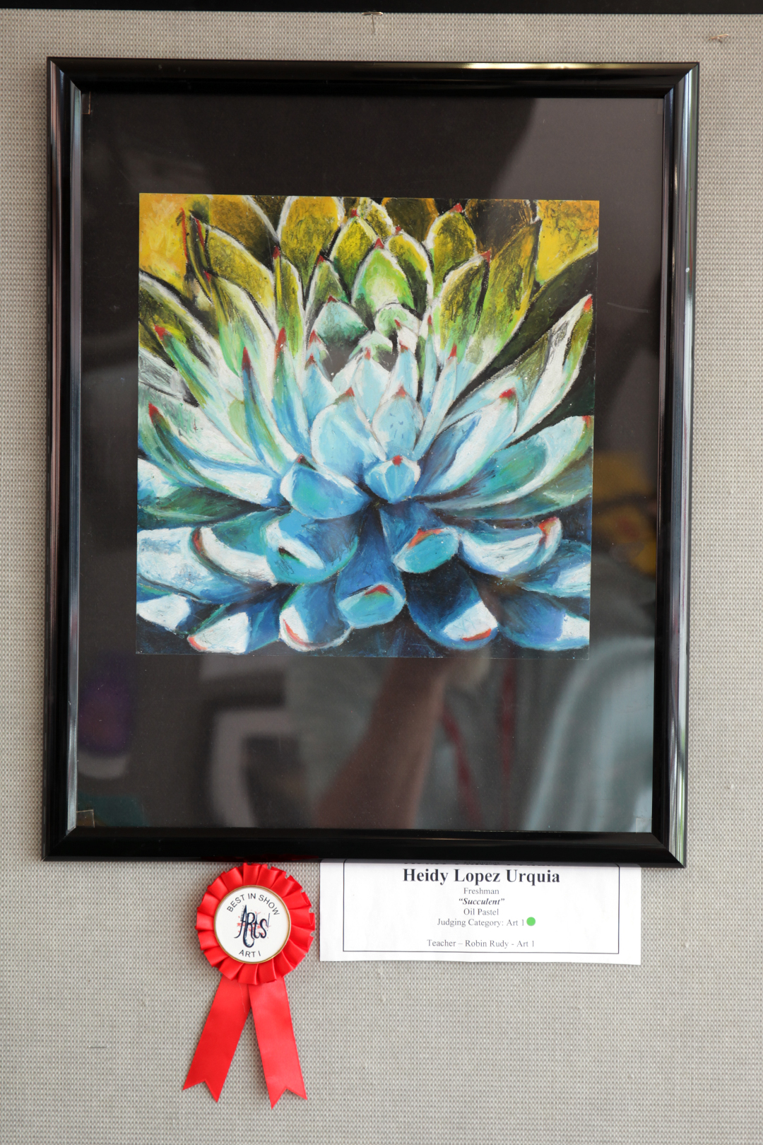 A framed painting of a succulent with blue and green leaves is displayed on a wall, with a red ribbon and a name tag below it.