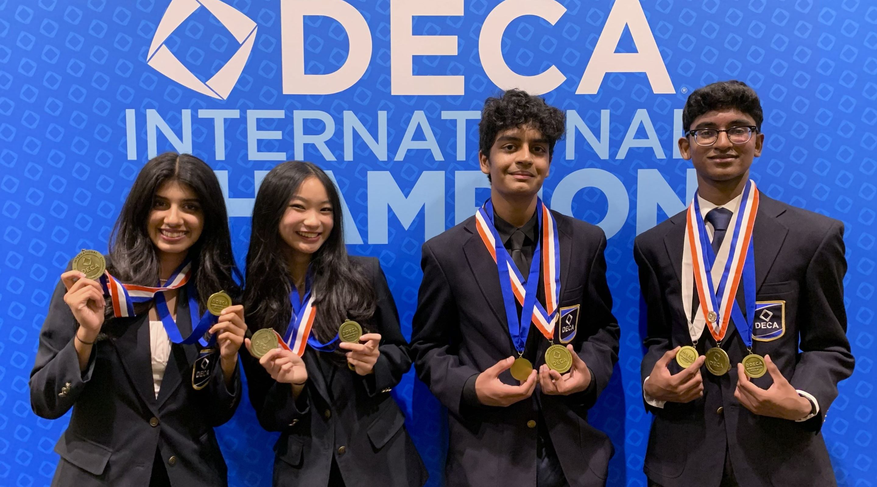 Four smiling students in blazers and medals stand in front of a blue DECA International Champion banner.