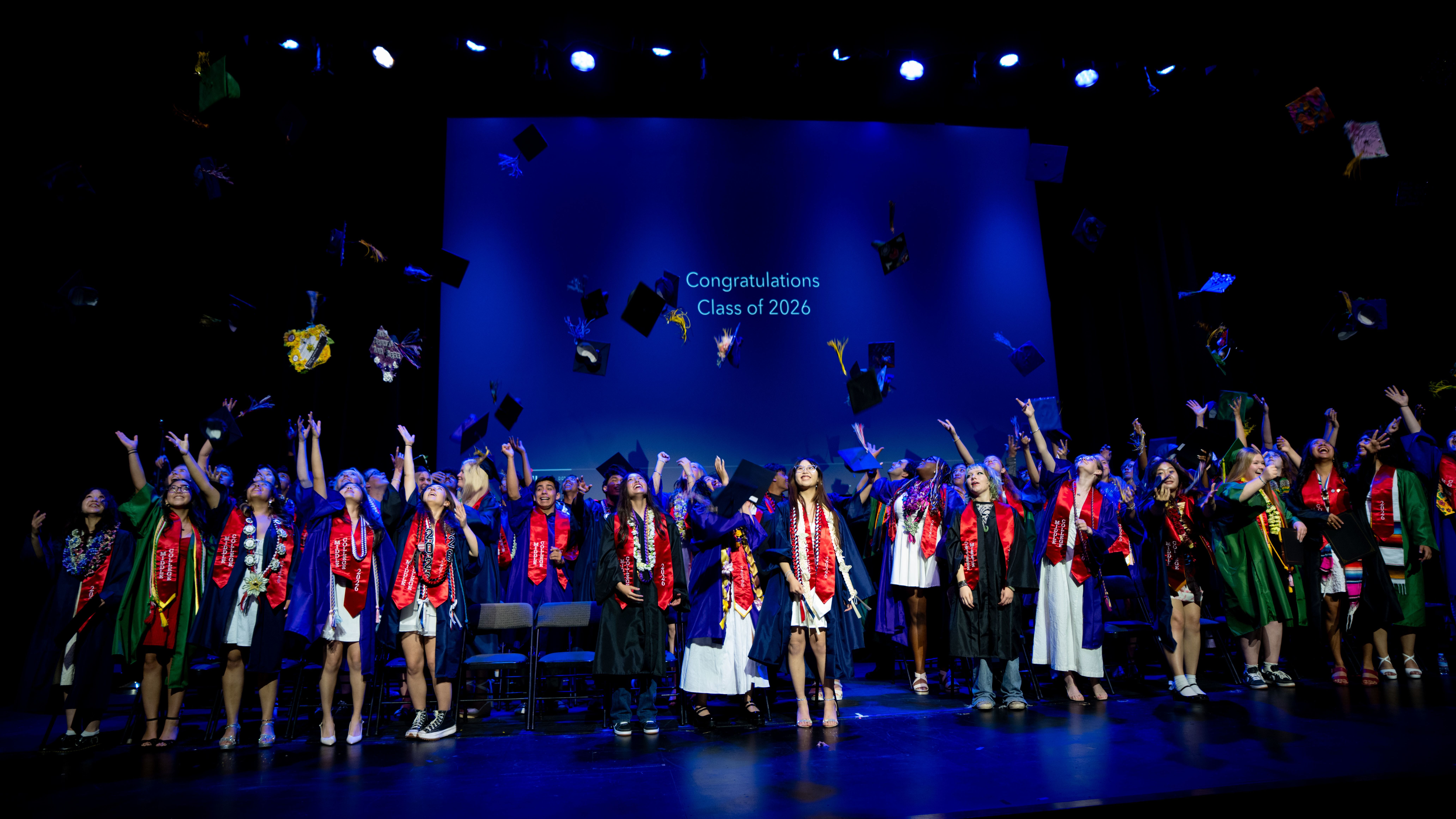 Middle College graduates in caps and gowns toss their hats in the air on a stage.