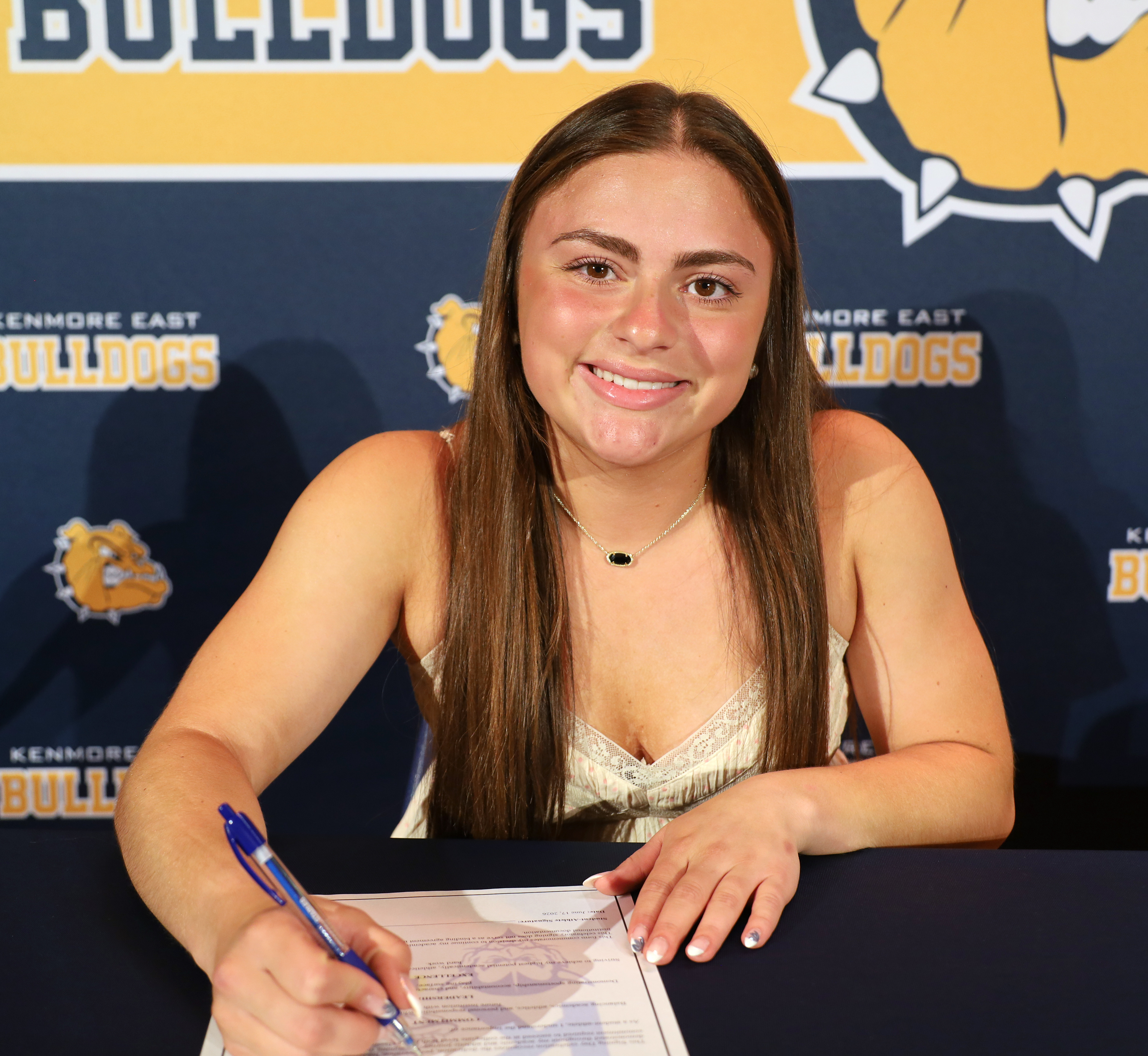 A smiling young woman in a white top signs a document at a table, with a "Kenmore East Bulldogs" banner in the background.