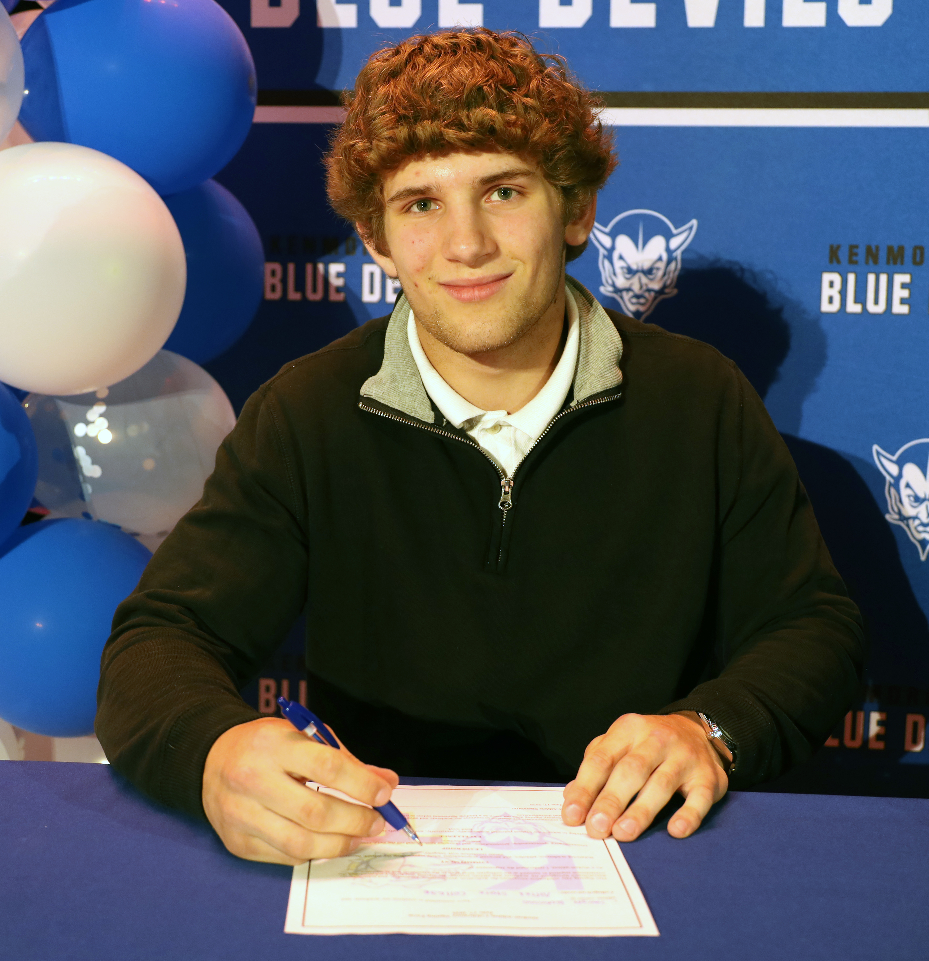 A young man with curly red hair signs a document at a table, with blue and white balloons and a "Blue Devils" banner in the background.