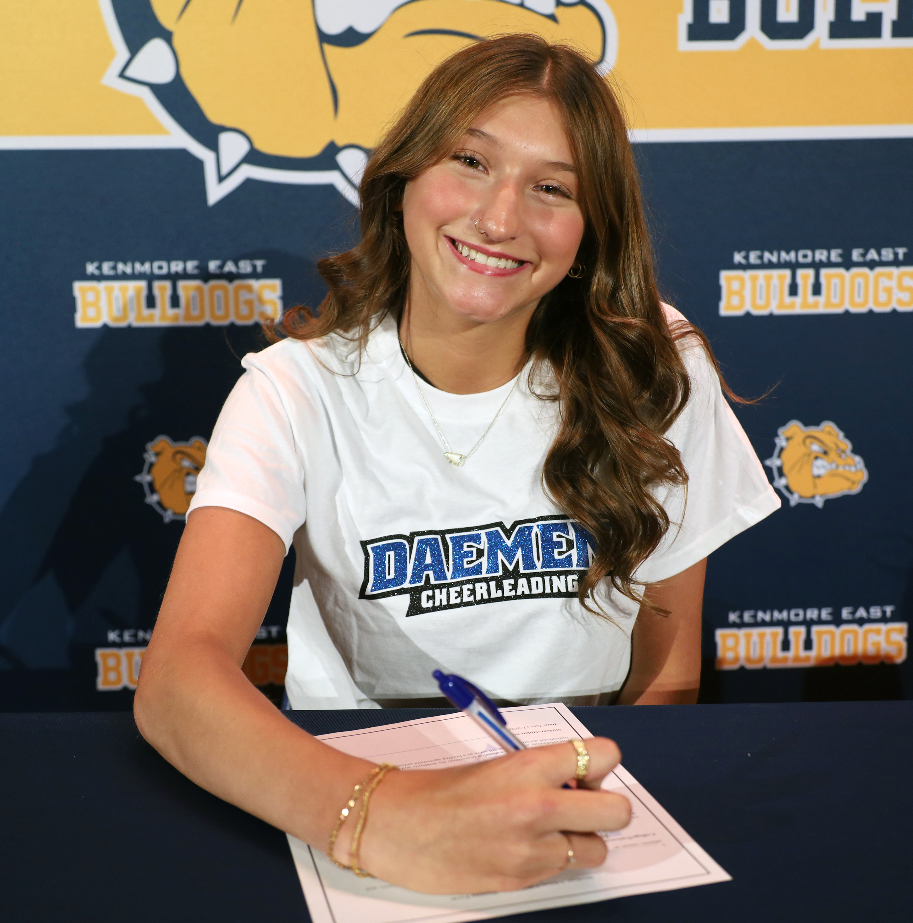 A smiling young woman in a Daemen Cheerleading shirt signs a document at a table, with a Kenmore East Bulldogs banner in the background.