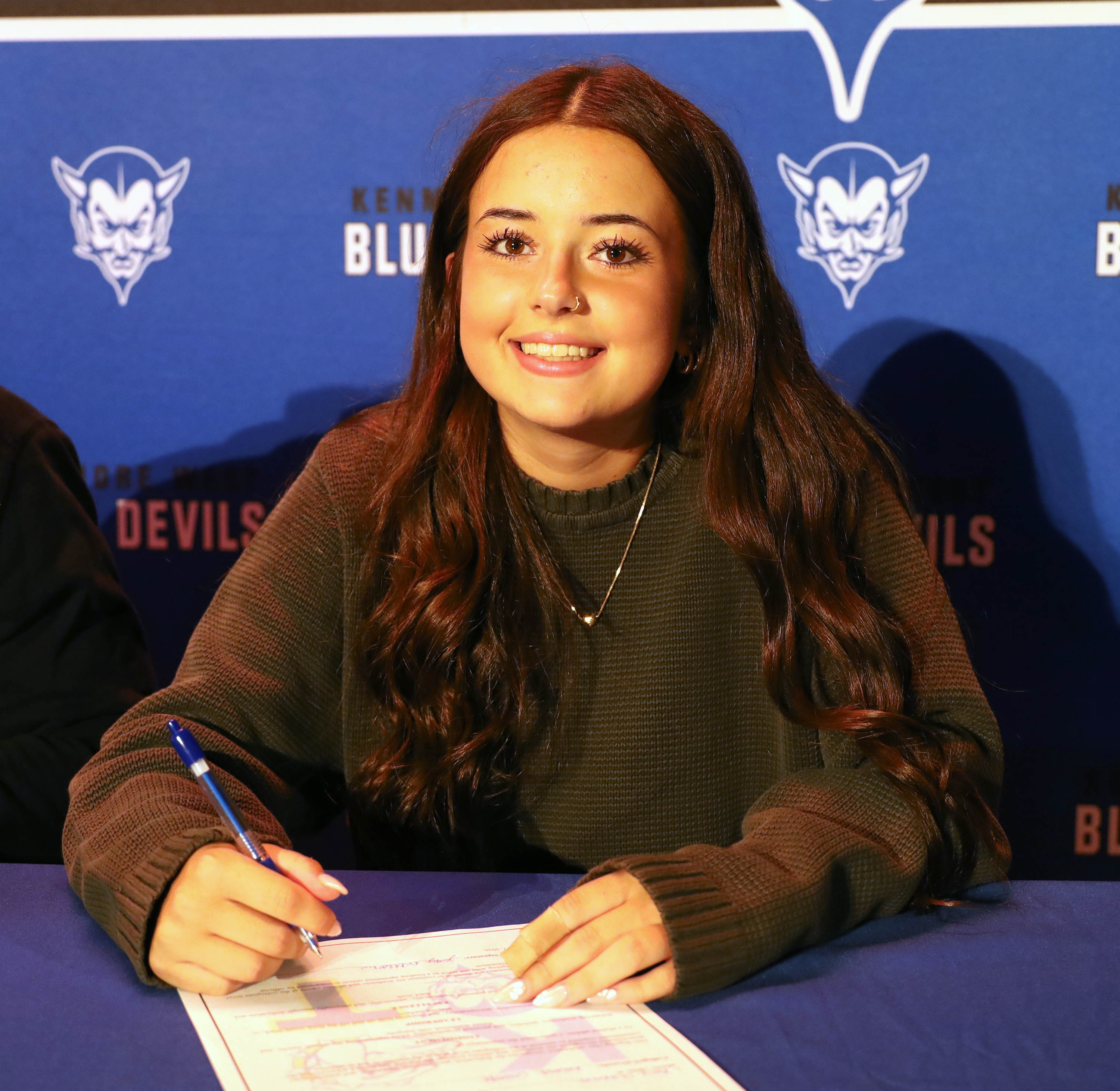 A young woman with long brown hair smiles as she signs a document at a table, with a blue banner featuring devil logos in the background.