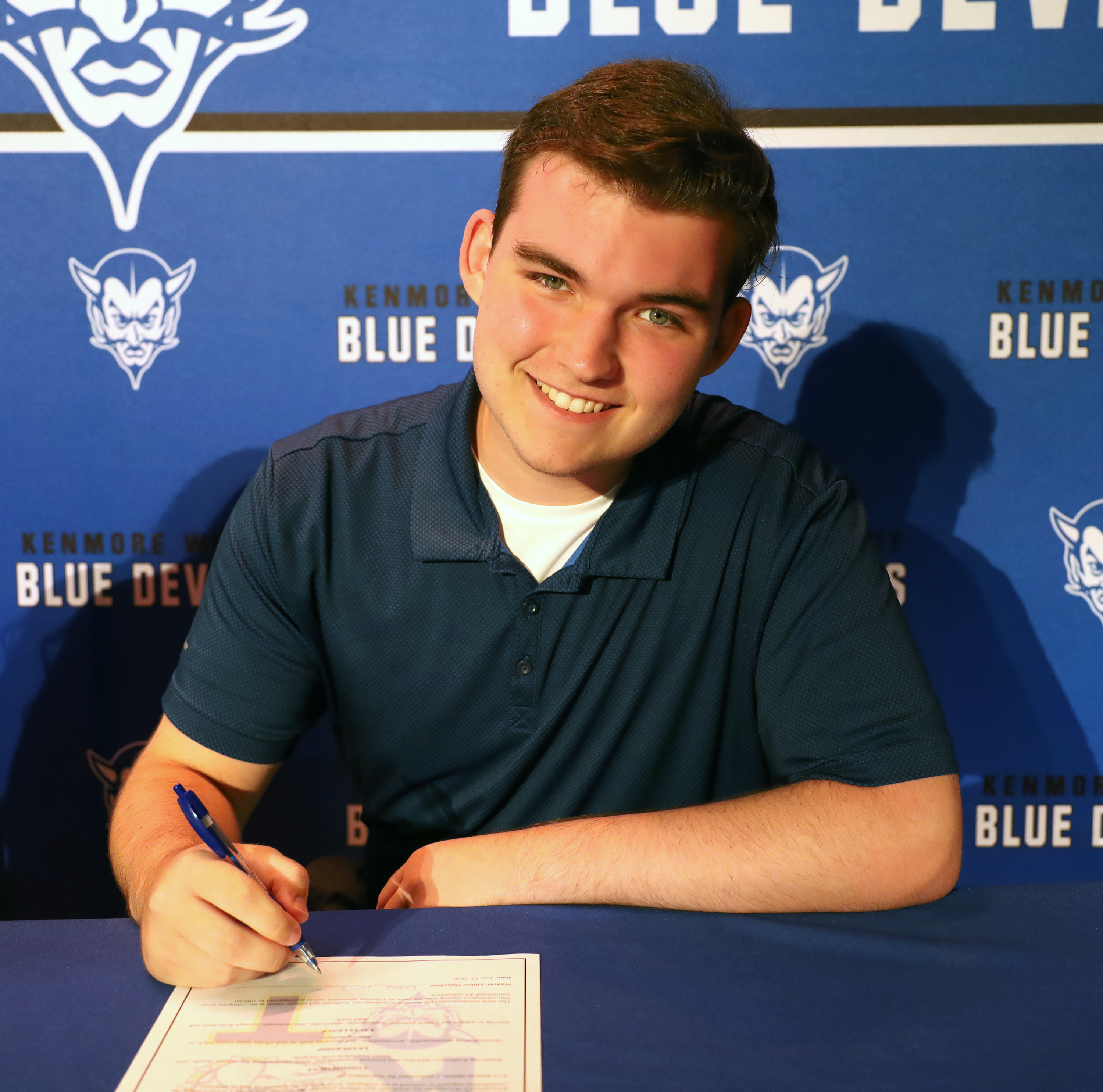 A smiling young man in a blue polo shirt signs a document at a table, with a blue banner featuring the Kenmore Blue Devils logo behind him.