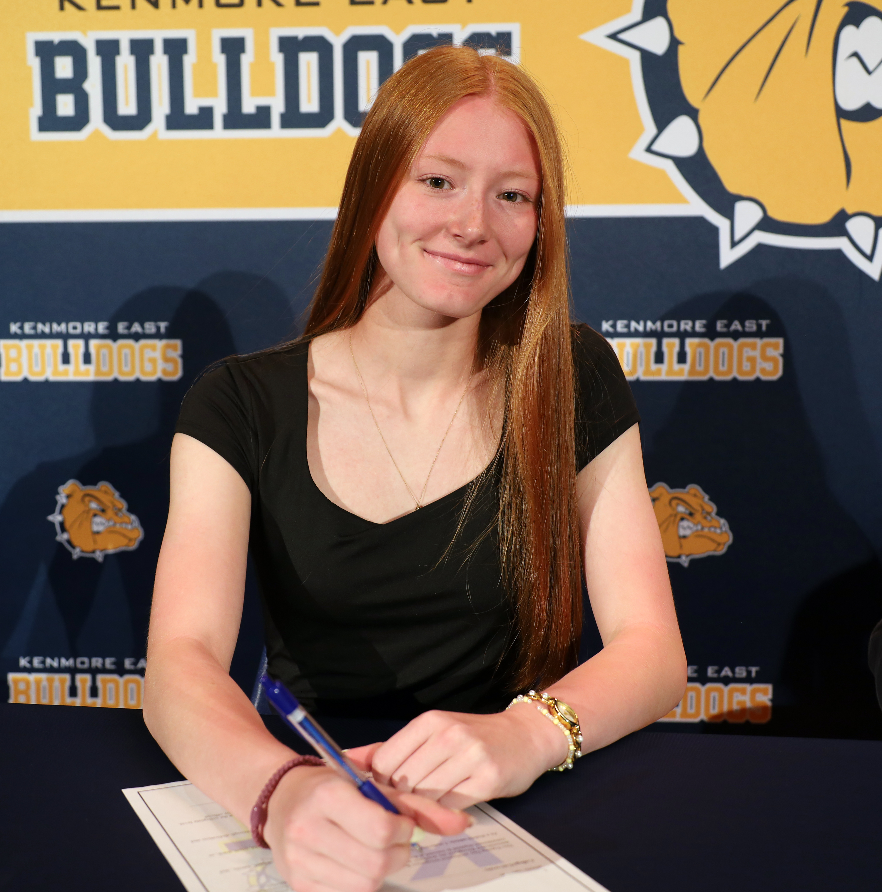 A young woman with long red hair smiles as she signs a document in the foreground, with a backdrop featuring the Kenmore East Bulldogs logo.