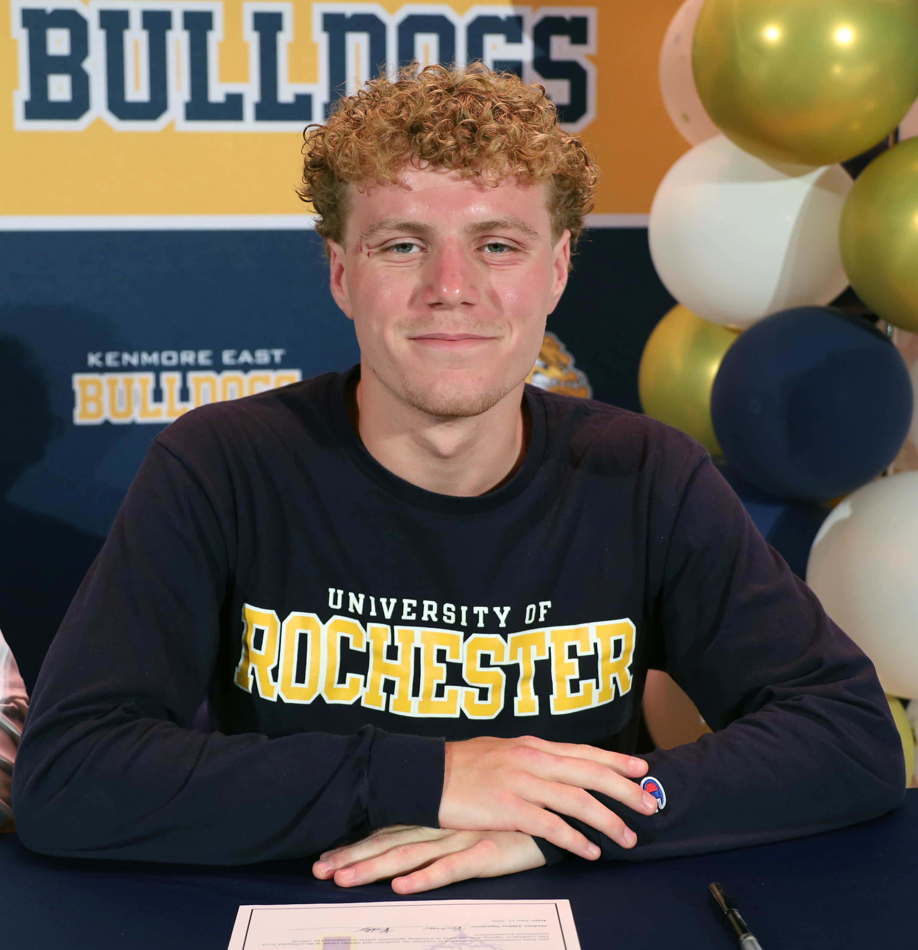 A young man with curly blonde hair, wearing a navy long-sleeved shirt with "University of Rochester" printed on it, sits at a table with a document and pen in front of him, while a backdrop displays "BULLDOGS" in large letters and a cluster of gold, white, and navy balloons.