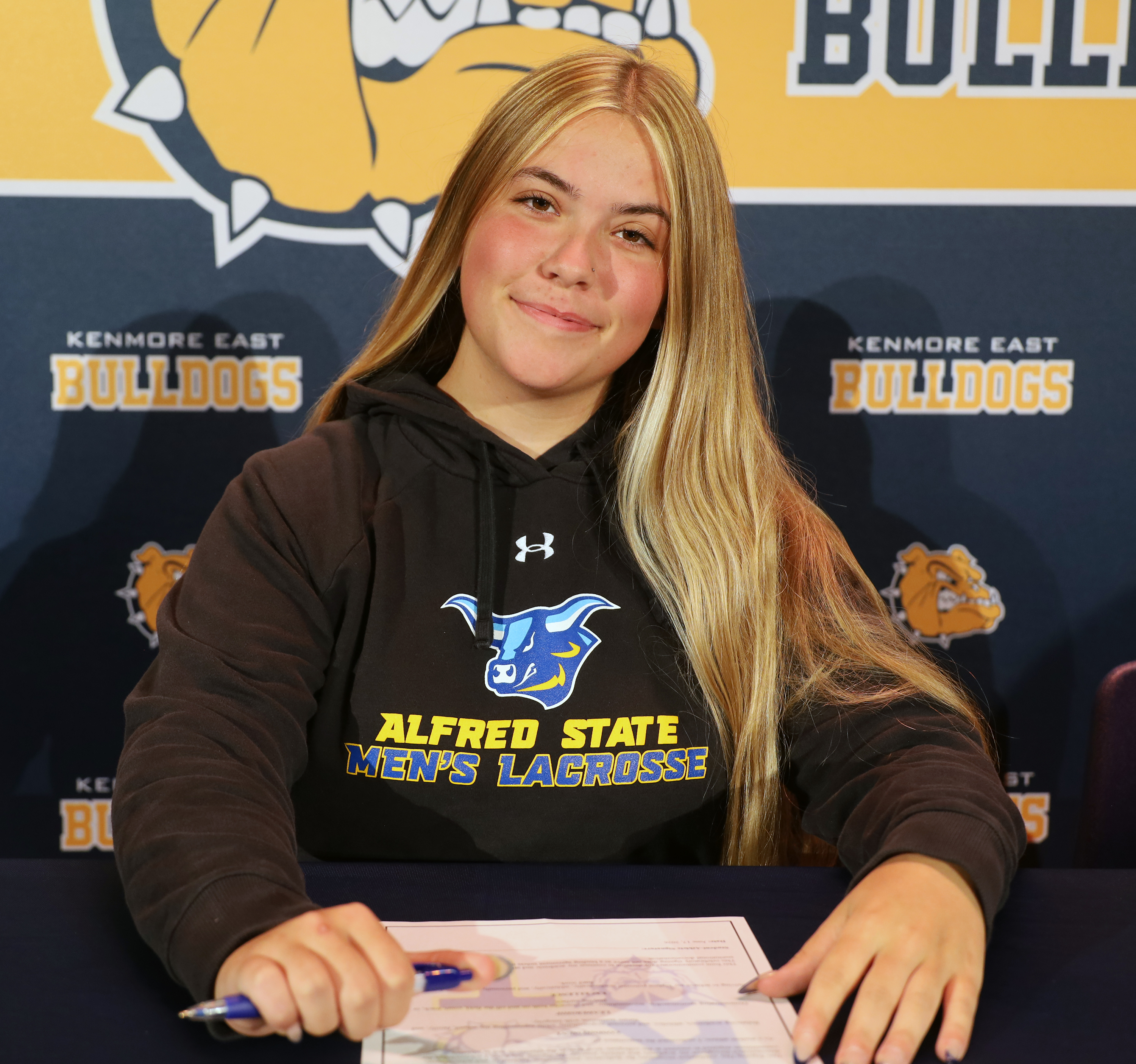 A young woman with long blonde hair smiles as she signs a document in front of a backdrop featuring the Kenmore East Bulldogs logo.