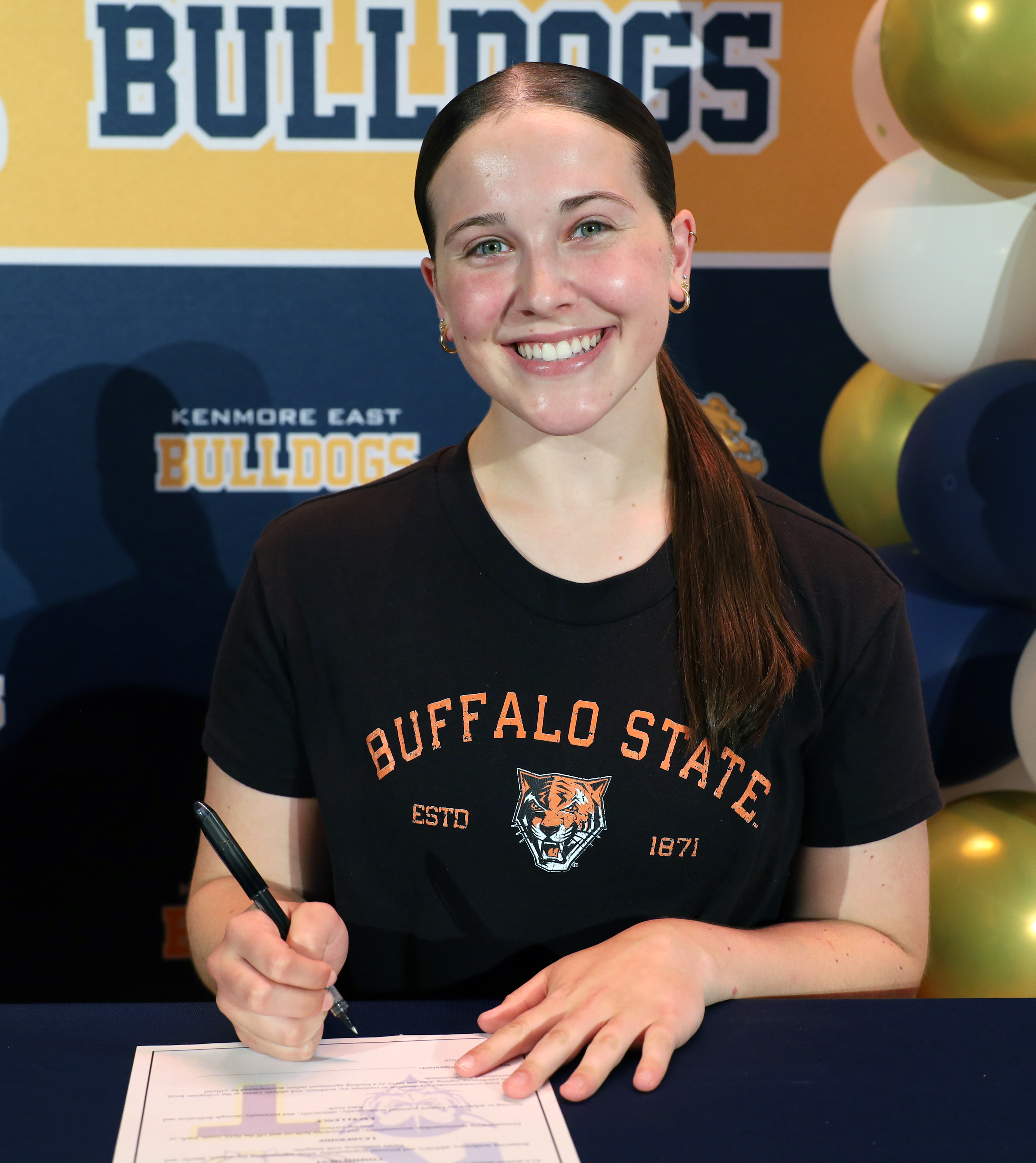 A smiling young woman in a black "Buffalo State" t-shirt signs a document in the foreground, with a "Bulldogs" banner and balloons in the background.