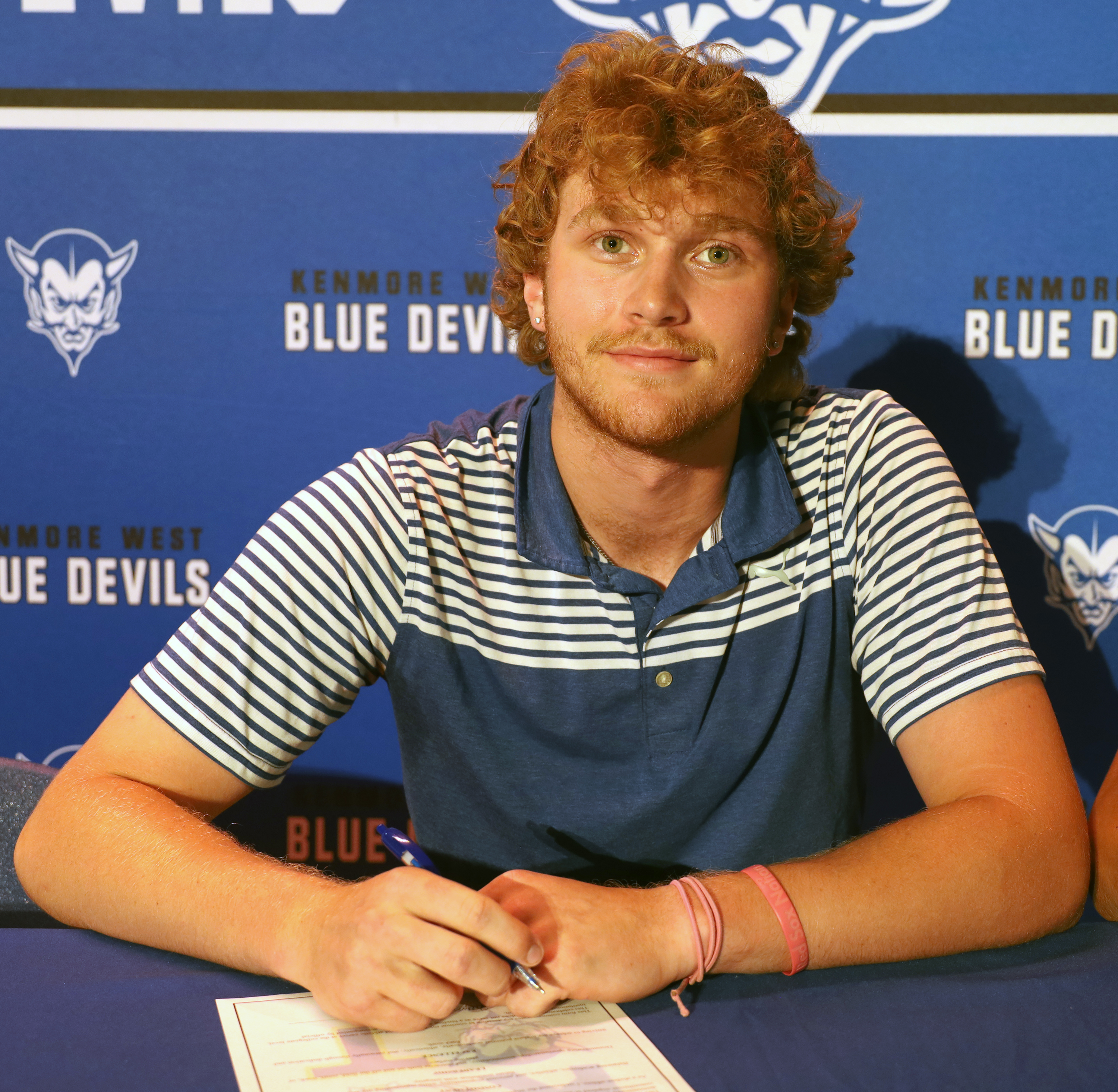A young man with red hair and a striped shirt sits at a table in front of a blue banner with "KENMORE WEST BLUE DEVILS" printed on it.