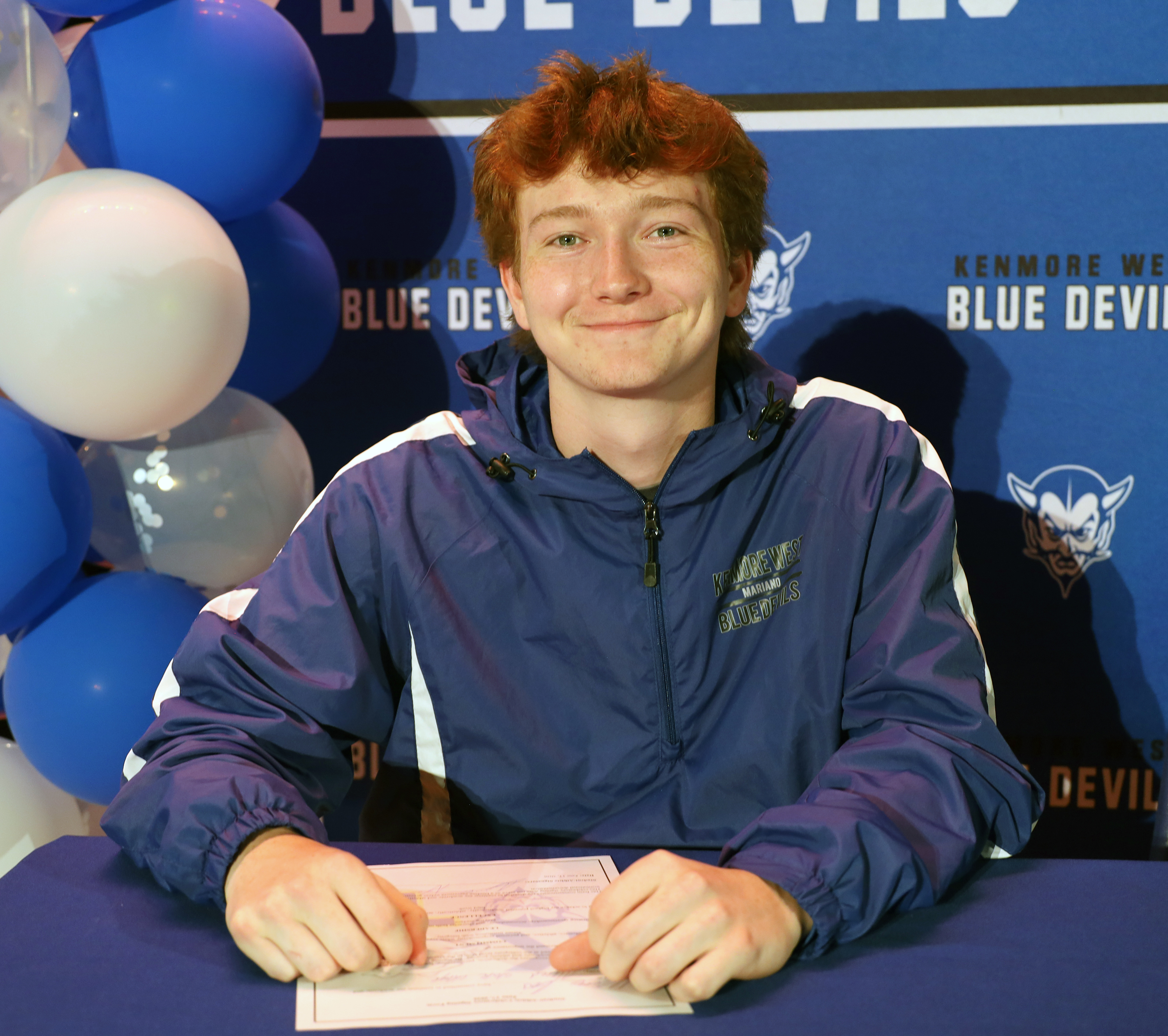A young man with red hair smiles as he sits at a table with a document in front of him, while blue and white balloons and a banner with "KENMORE WEST BLUE DEVILS" are in the background.