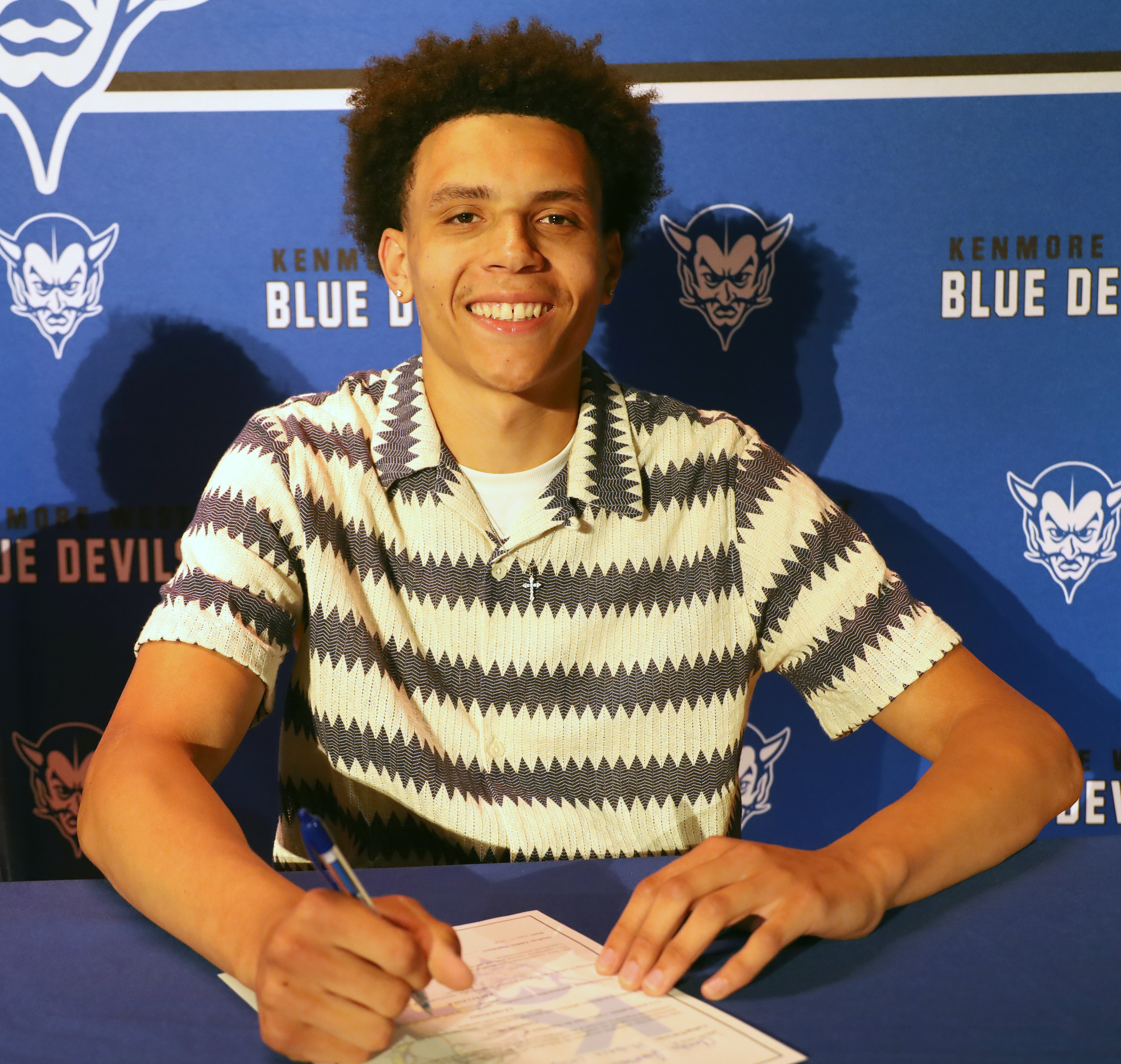A young man with curly hair smiles as he signs a document at a table, with a blue banner featuring the Kenmore Blue Devils logo behind him.
