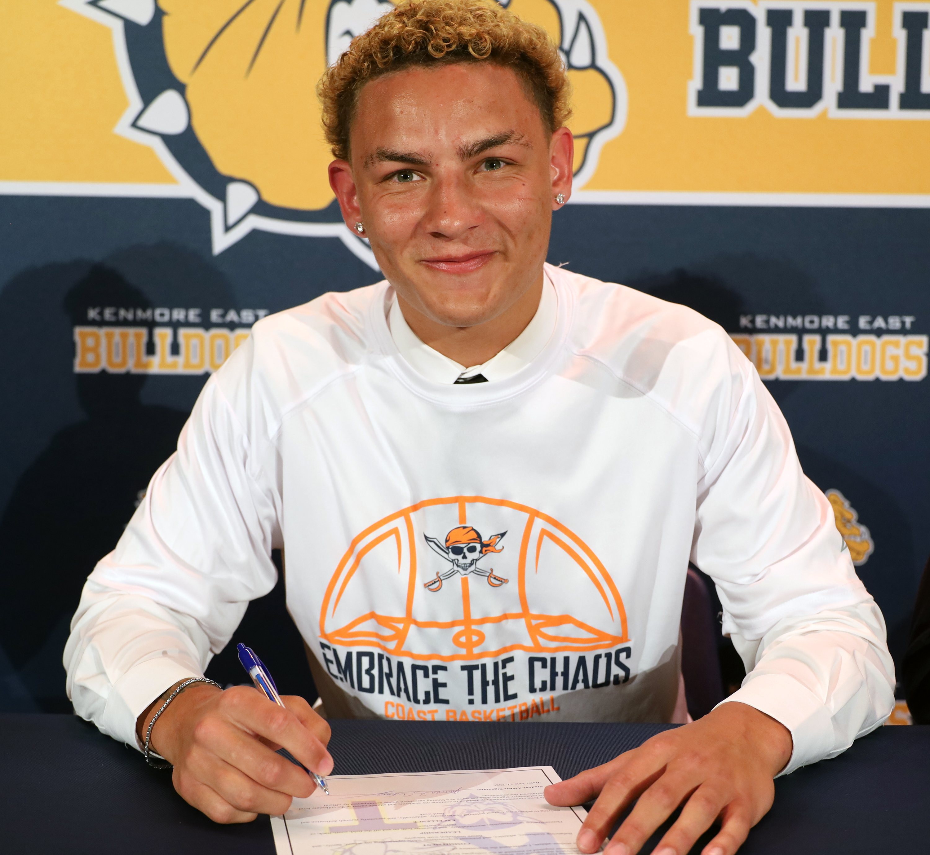 A young man with curly blonde hair smiles as he signs a document at a table, with a "Bulldogs" banner in the background.