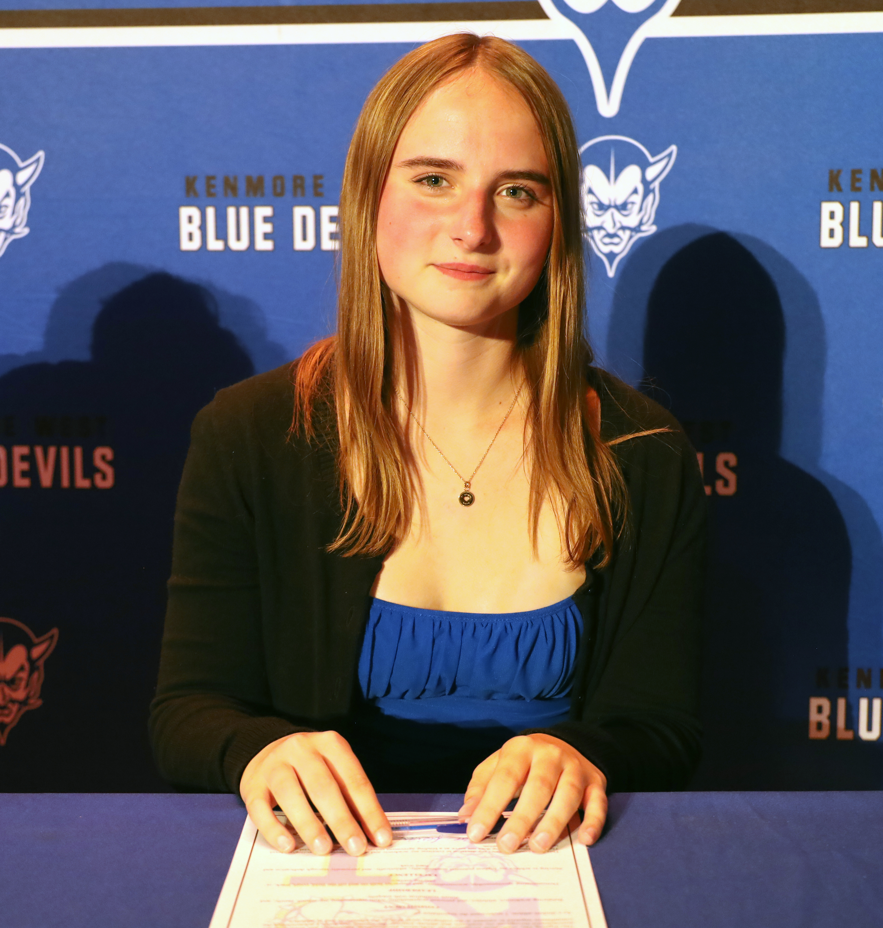 A young woman with long blonde hair sits at a table signing a document, with a blue banner featuring the "Kenmore Blue Devils" logo behind her.