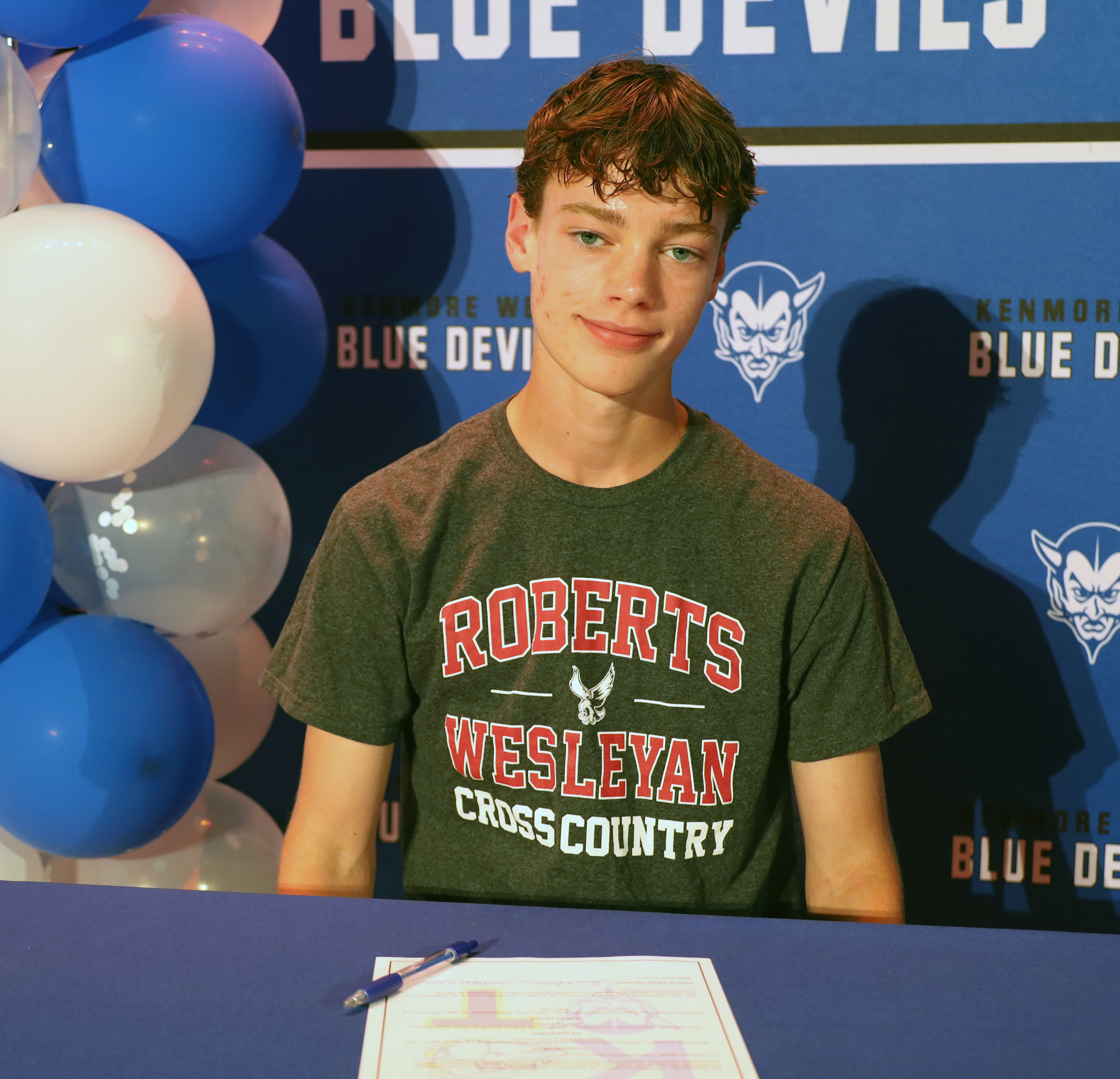 A young man in a "Roberts Wesleyan Cross Country" t-shirt smiles in front of a blue backdrop with "Blue Devils" logos and a cluster of blue and white balloons.