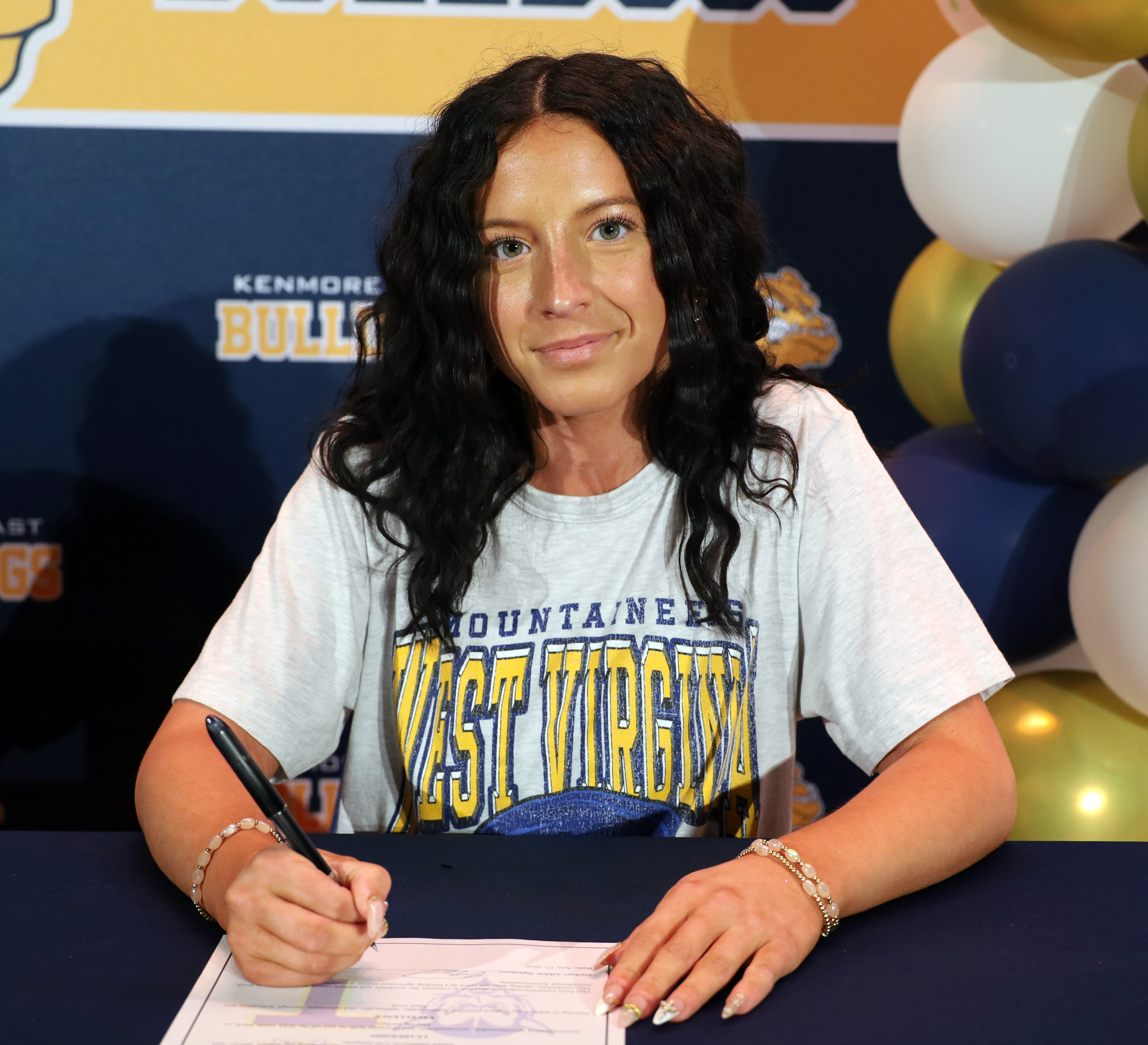 A woman in a "West Virginia Mountaineers" t-shirt signs a document at a table, with a "Kenmore Bulldogs" banner and balloons in the background.