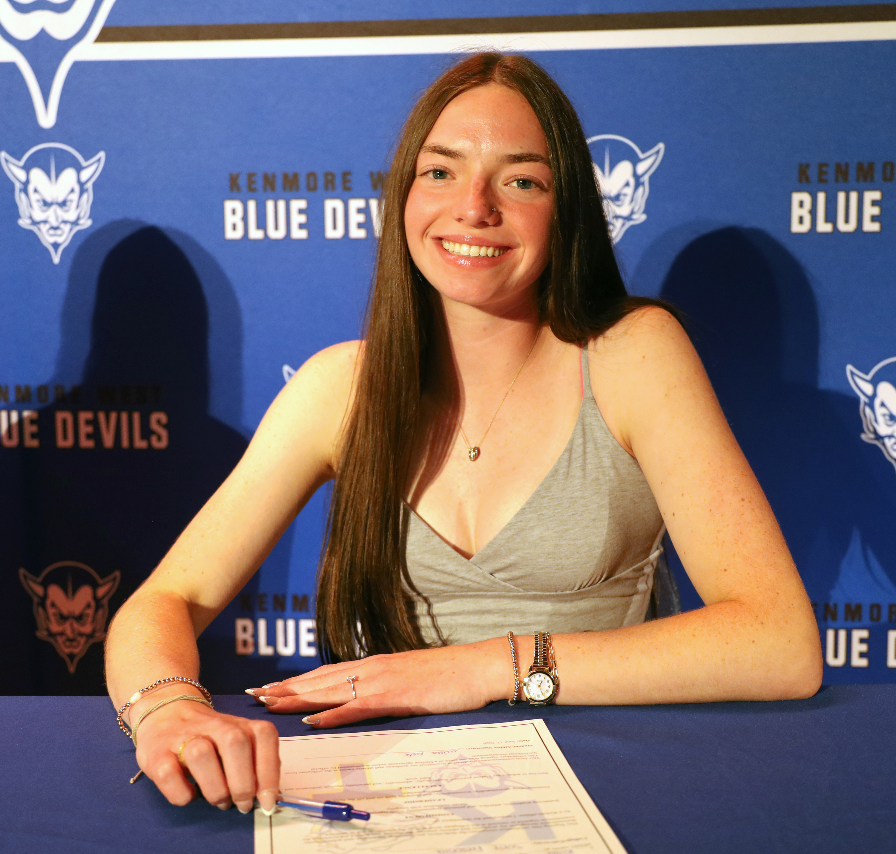 A smiling young woman in a grey tank top signs a document at a table, with a blue backdrop featuring the "Kenmore West Blue Devils" logo.