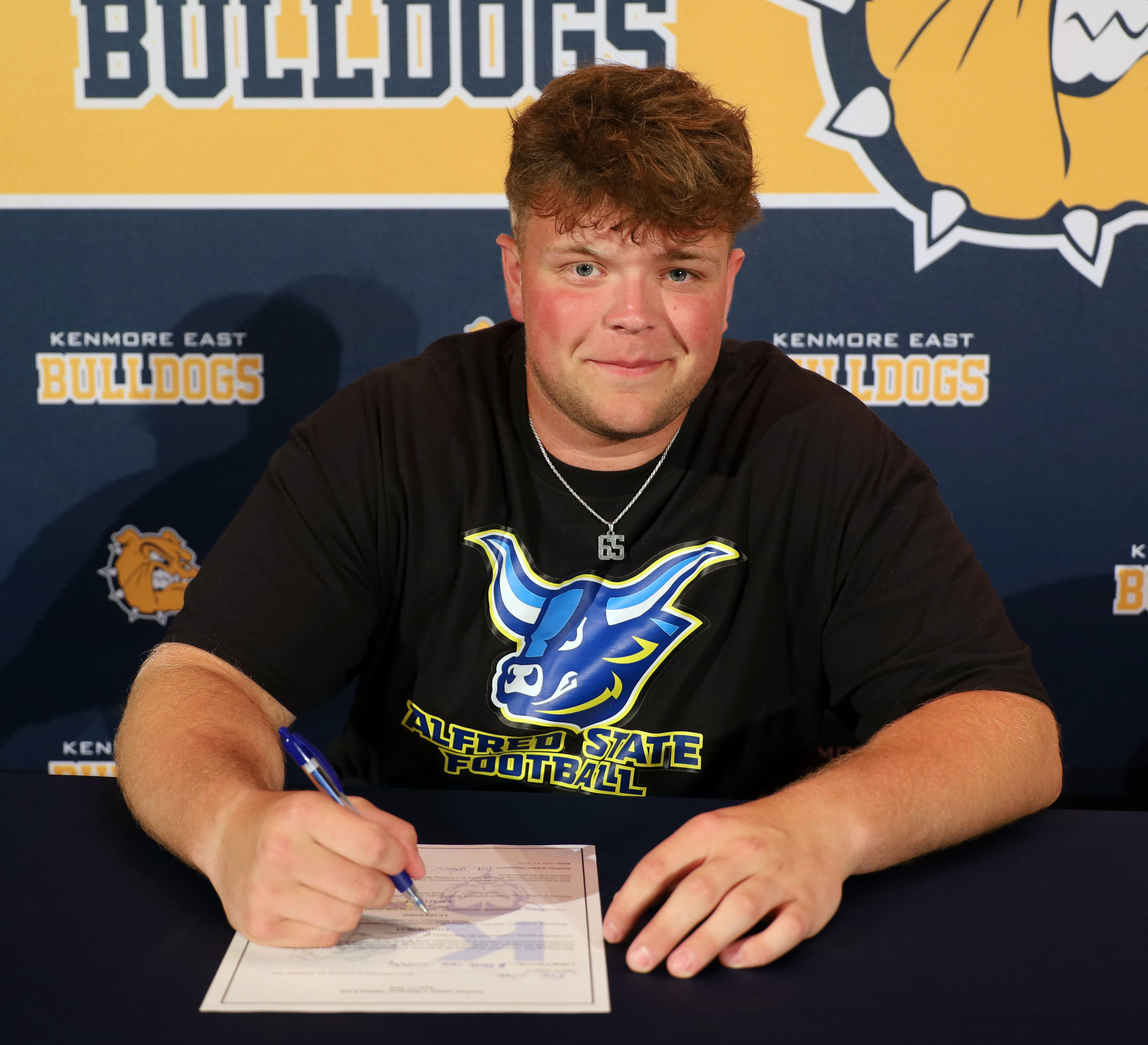 A young man in a black t-shirt with a blue bull logo signs a document at a table, with a backdrop featuring "BULLDOGS" lettering and a bulldog mascot.