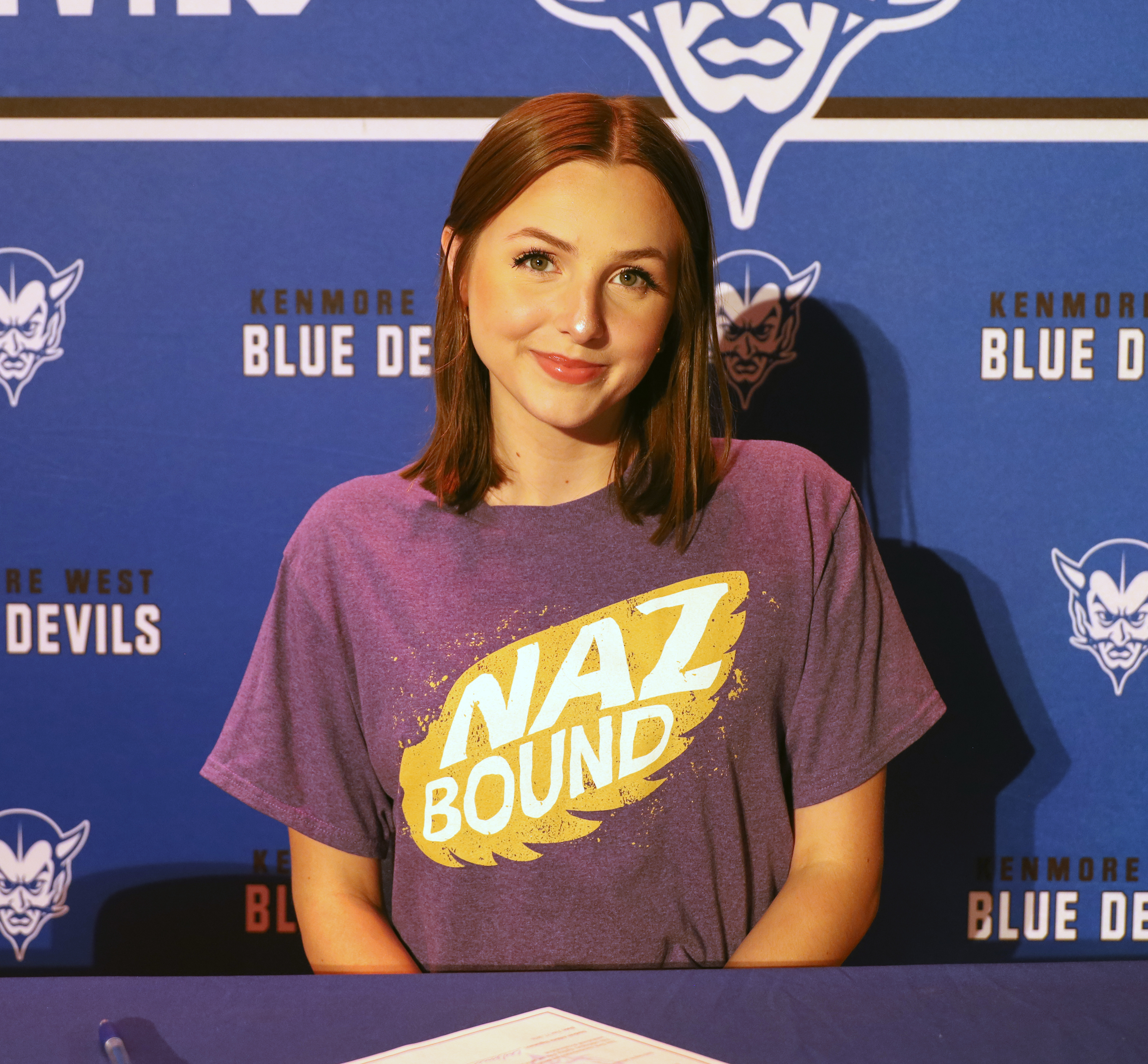 A young woman in a purple "NAZ BOUND" t-shirt smiles in front of a blue banner with "KENMORE BLUE DEVILS" printed on it.