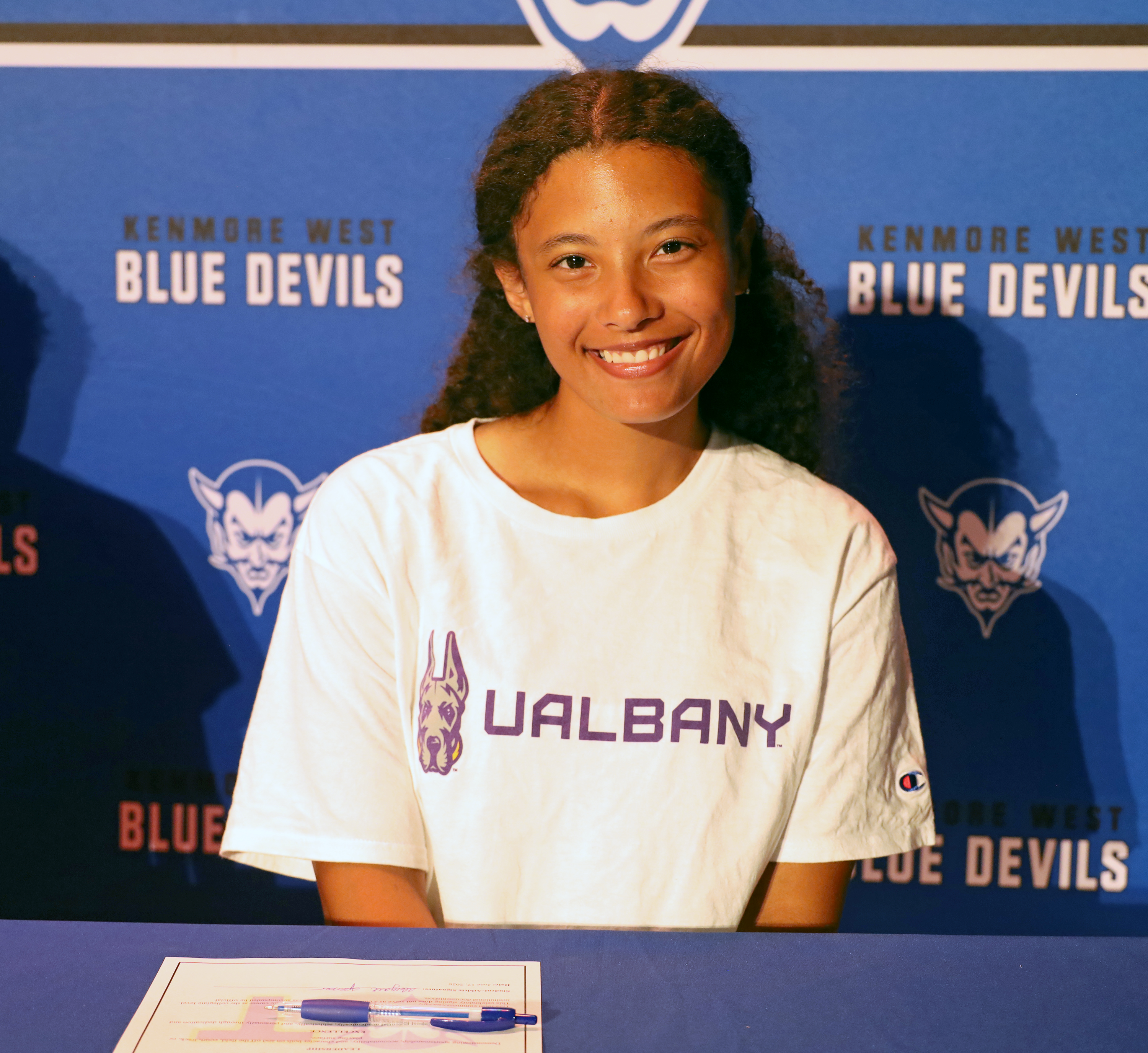 A smiling young woman in a "UALBANY" t-shirt sits at a table with a pen and paper in front of a blue backdrop featuring "KENMORE WEST BLUE DEVILS" logos.