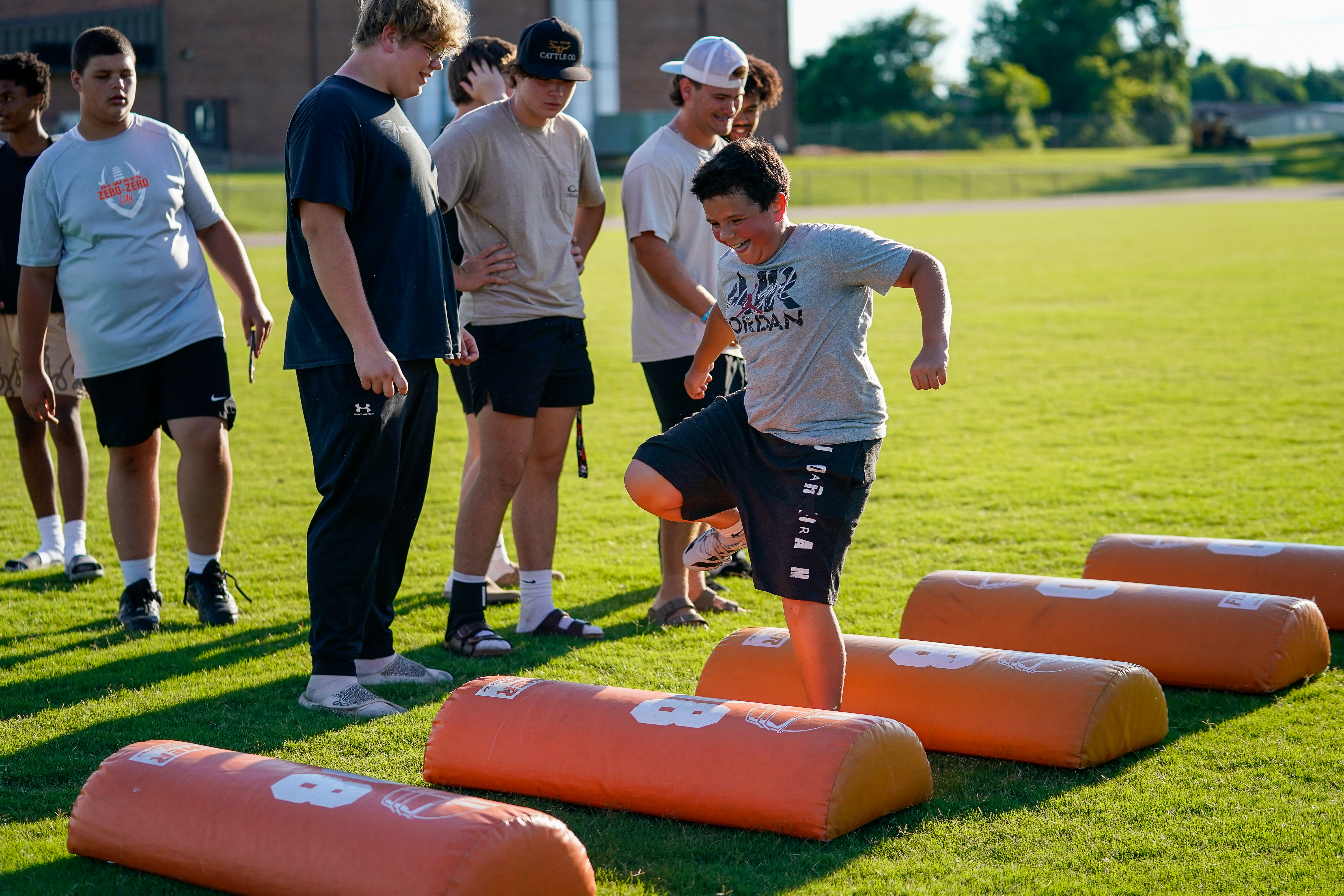 A young boy in the foreground jumps over orange blocking pads while a group of older boys watches in the background on a grassy field.
