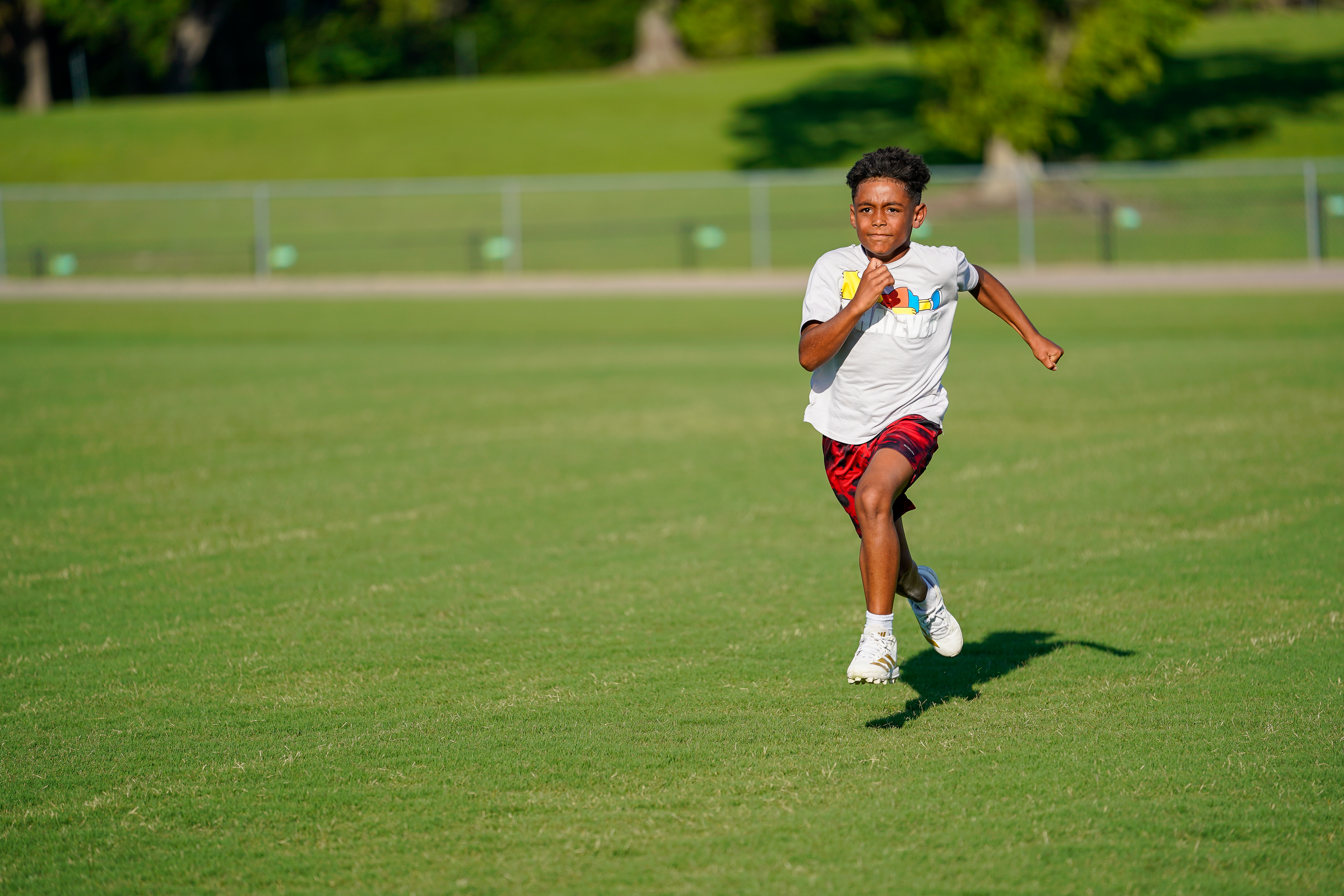 A young boy in athletic wear runs across a green field with trees and a fence in the background.