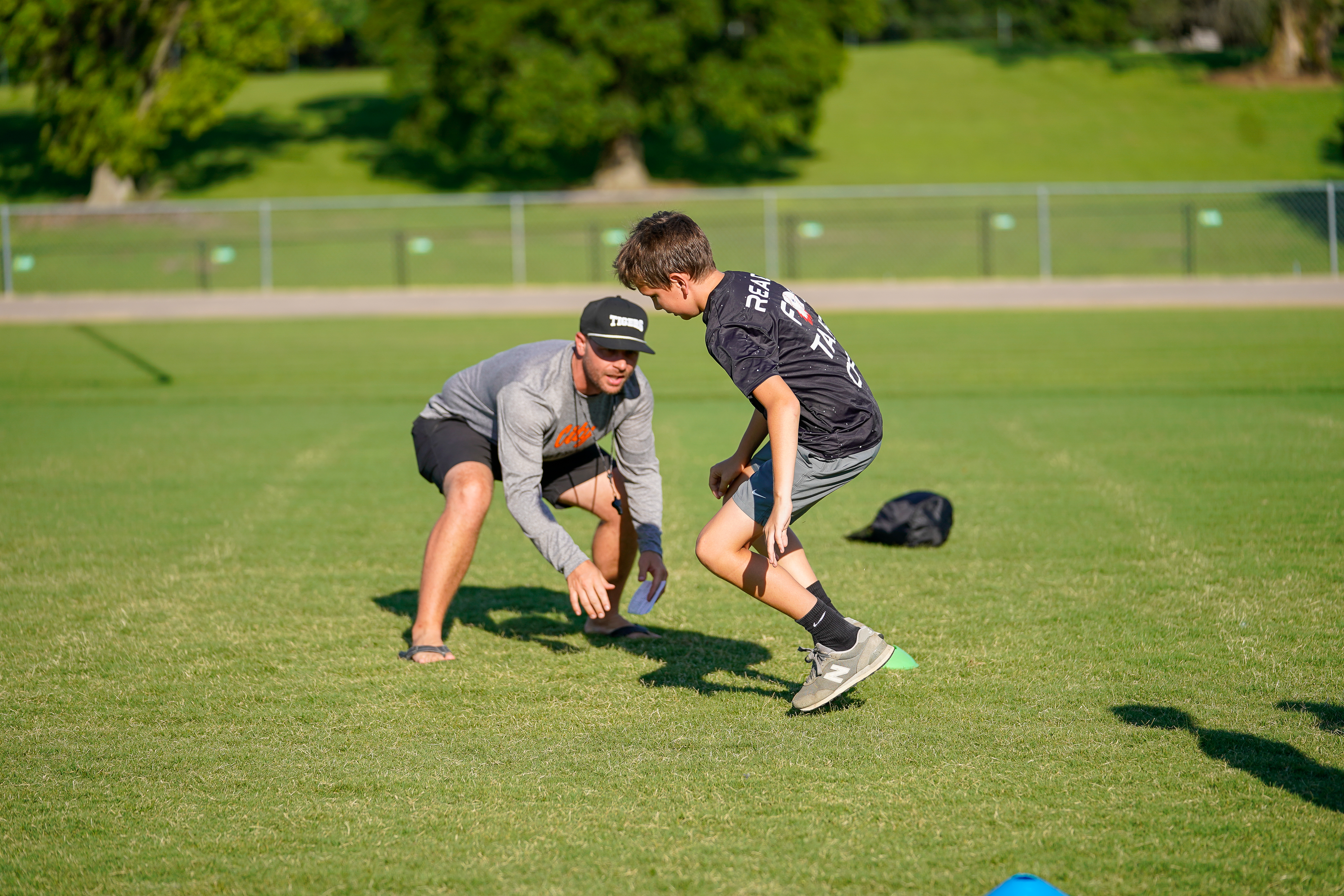 A coach instructs a young athlete on a grassy field, with a fence and trees in the background.
