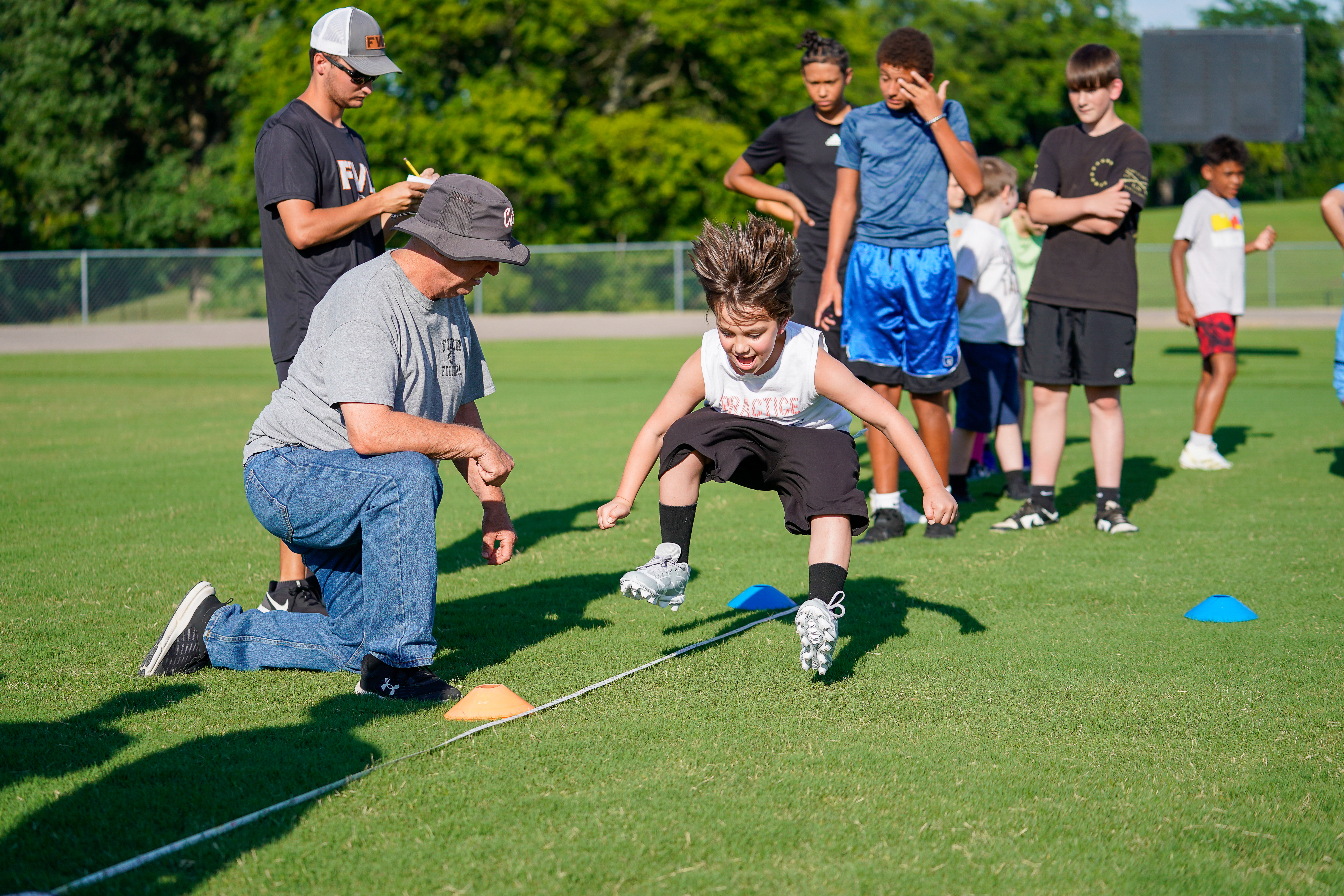 A young boy in athletic wear jumps over a rope on a grassy field while a coach watches, with other children and trees in the background.
