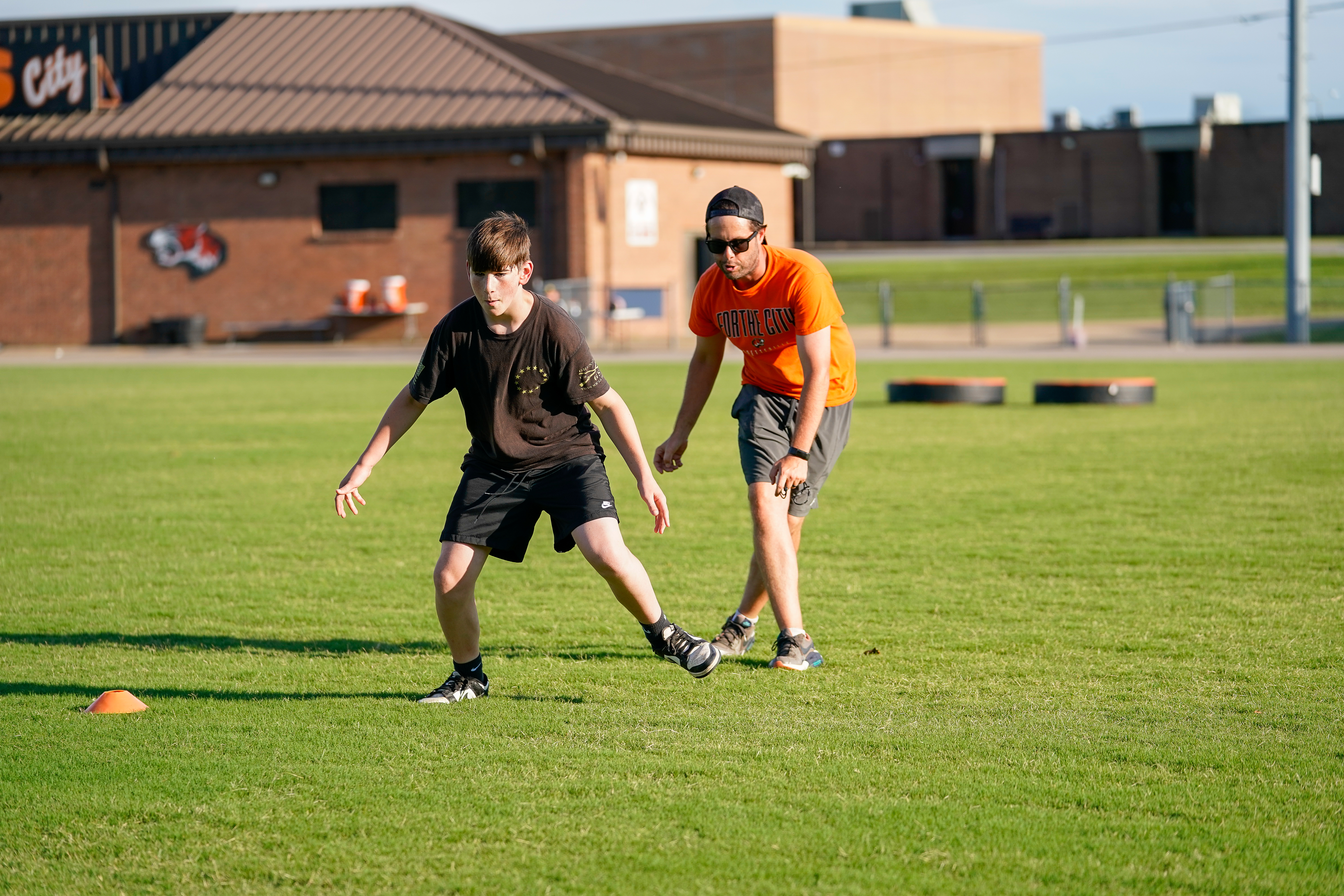 A young man in a black shirt and shorts practices drills on a grassy field with a coach in an orange shirt and sunglasses behind him, while a brick building stands in the background.