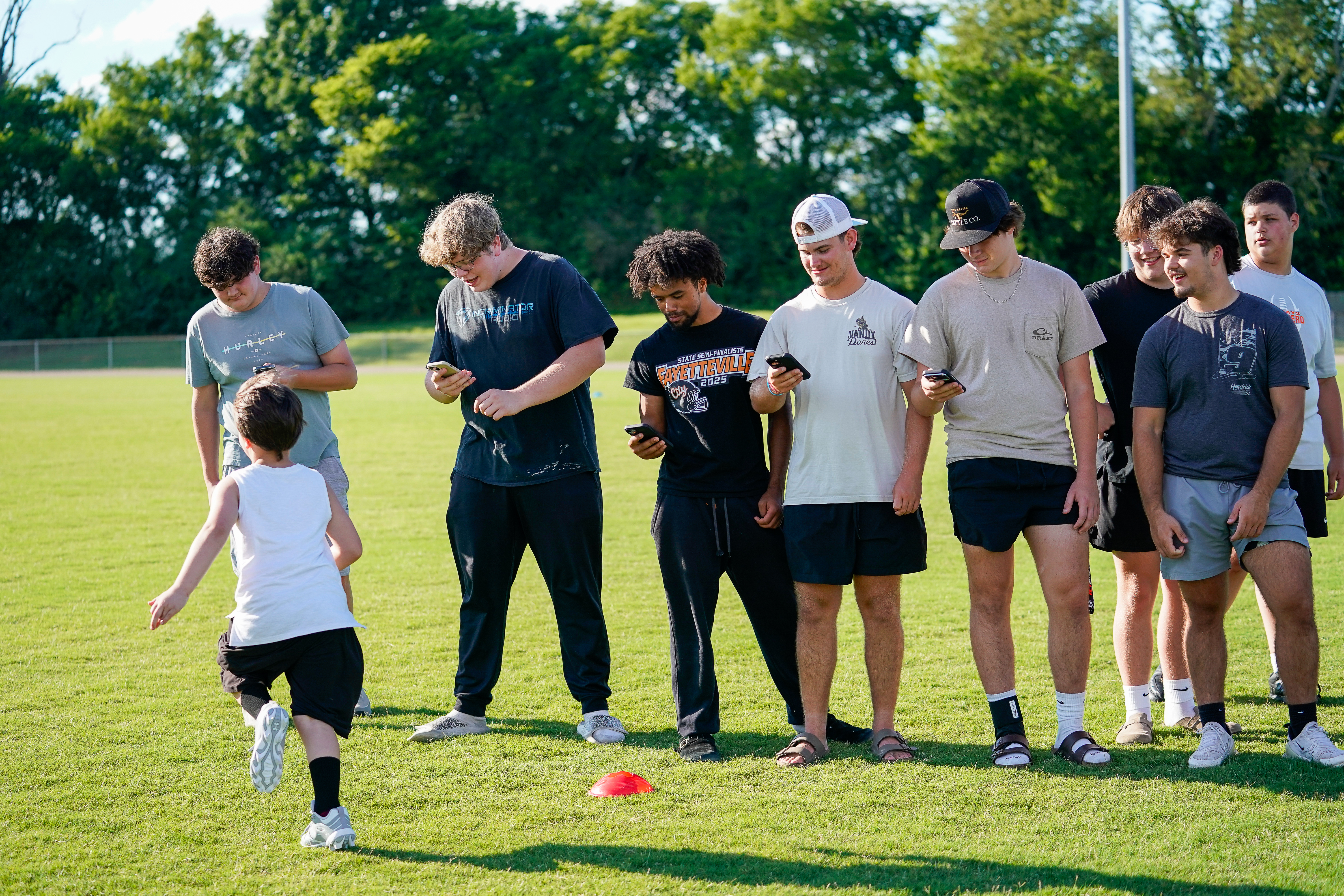A young boy in the foreground runs on a grassy field while a group of teenage boys in the background stand looking at their phones.