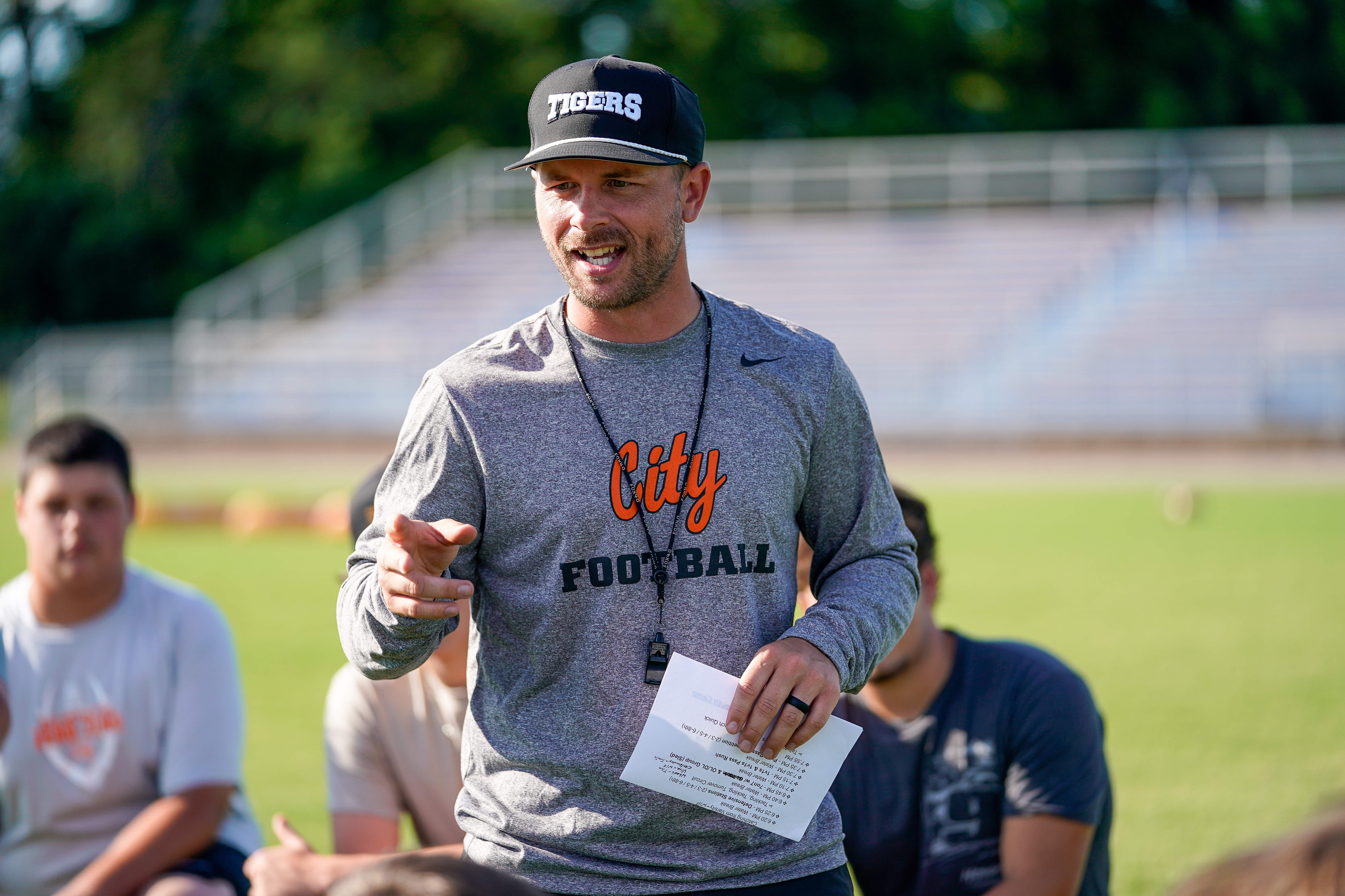 A man in a "Tigers" baseball cap and a gray "City Football" long-sleeved shirt gestures while speaking to a group of young men on a grassy field with bleachers in the background.