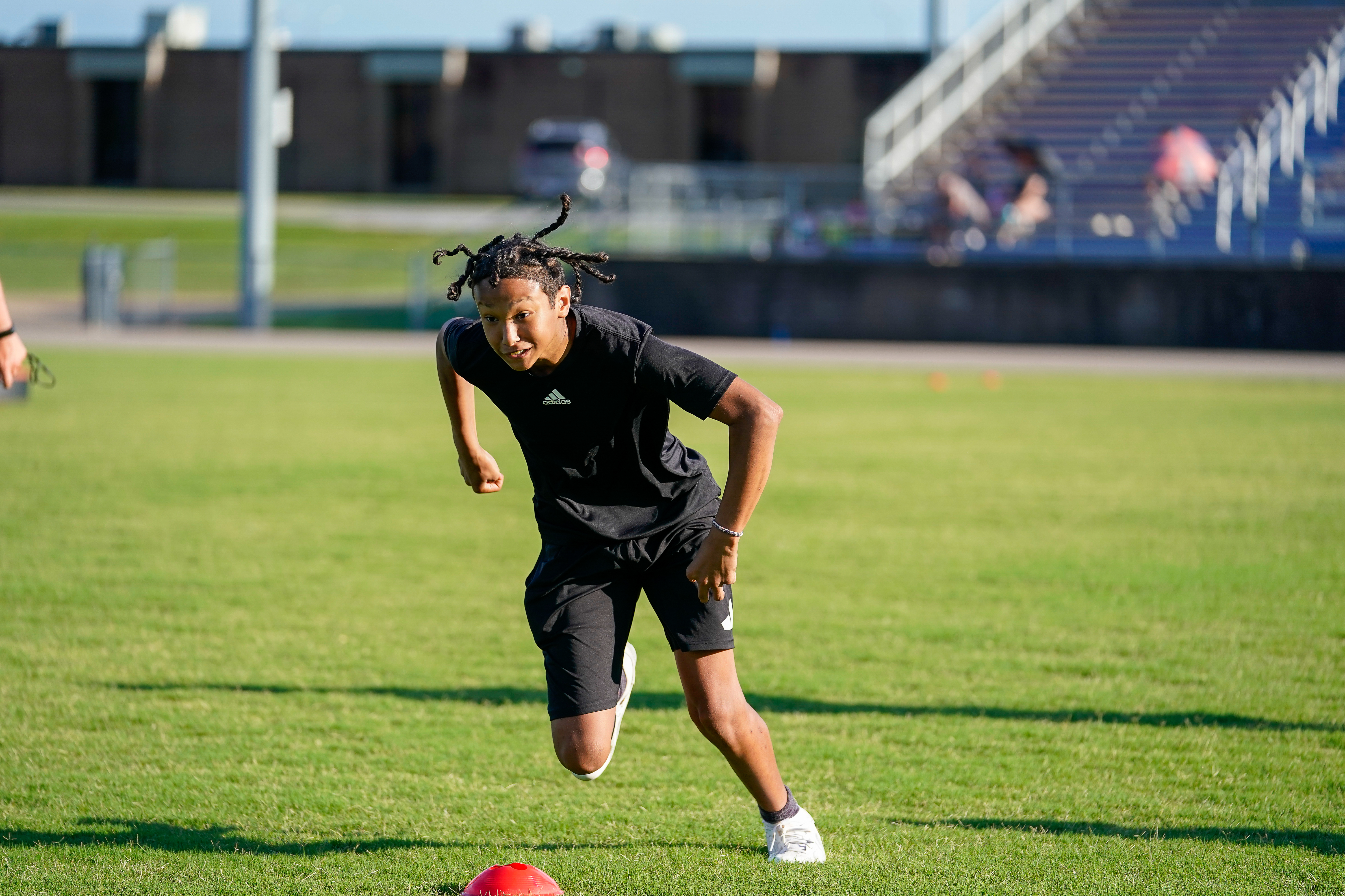 A young man in athletic wear sprints across a grassy field with stadium bleachers in the background.