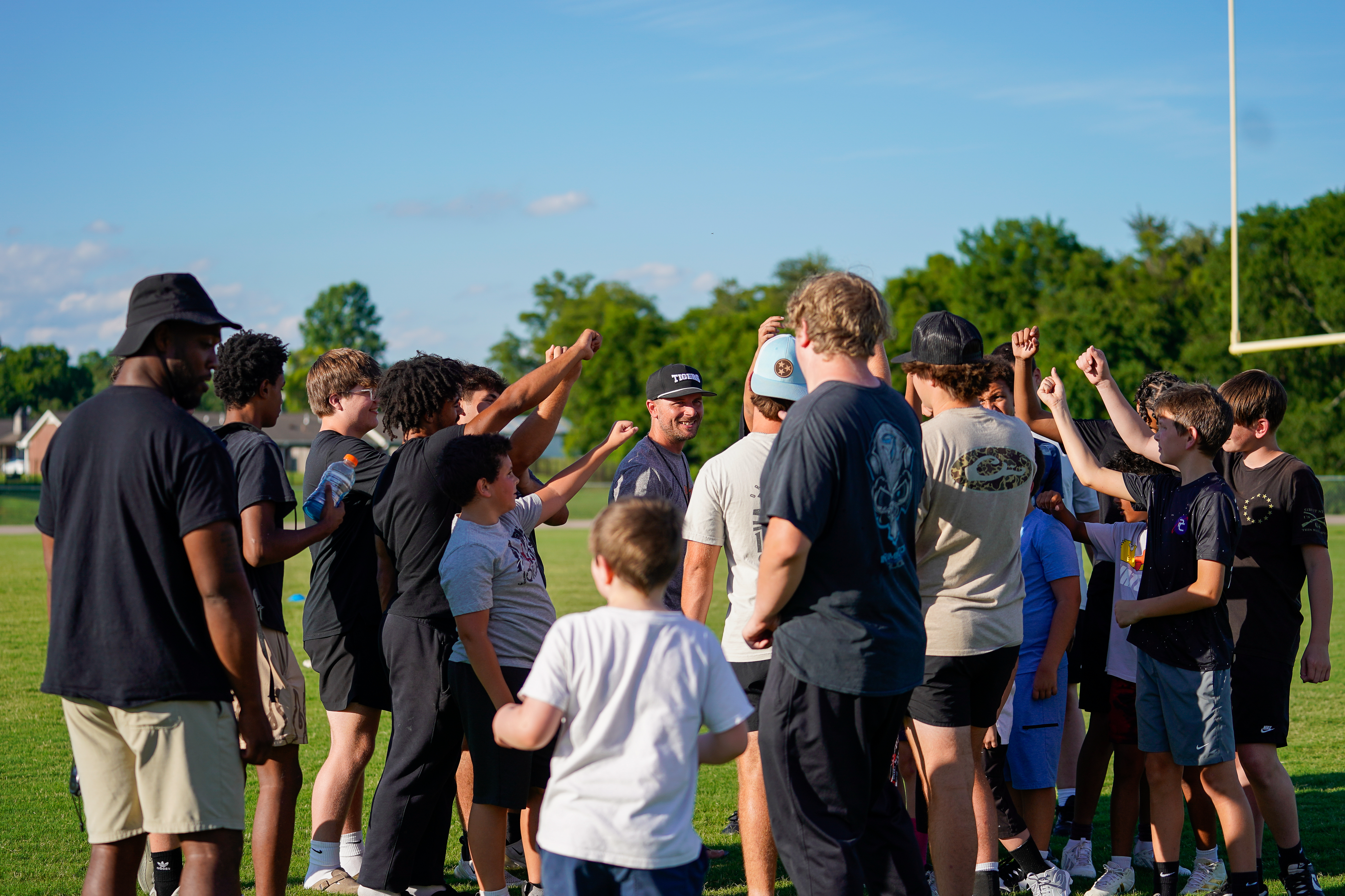 A group of young athletes huddle together on a grassy field with trees and a goalpost in the background.