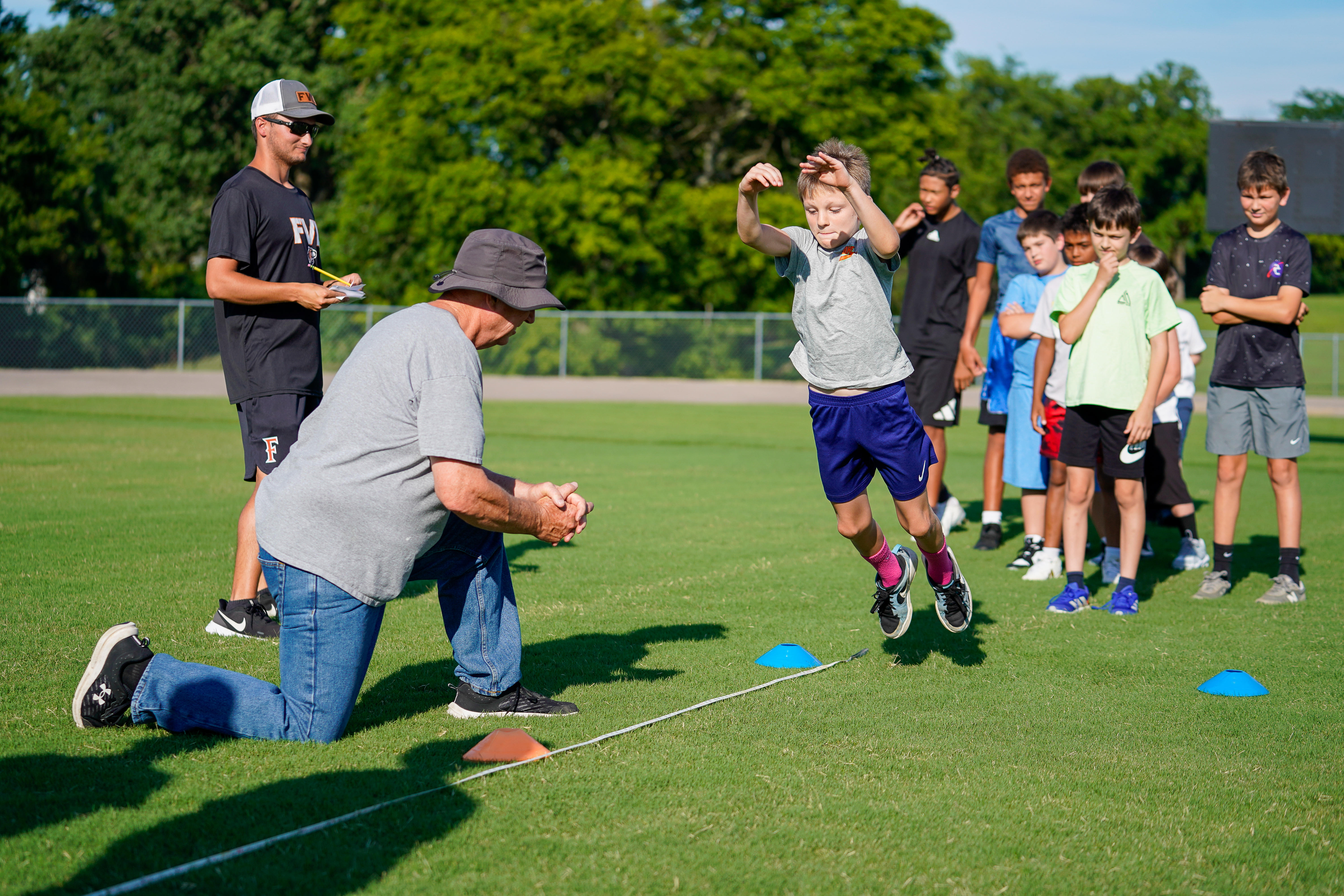 A young boy leaps over cones on a grassy field while a man kneels to observe, with a group of other boys and a coach standing in the background under a clear sky.