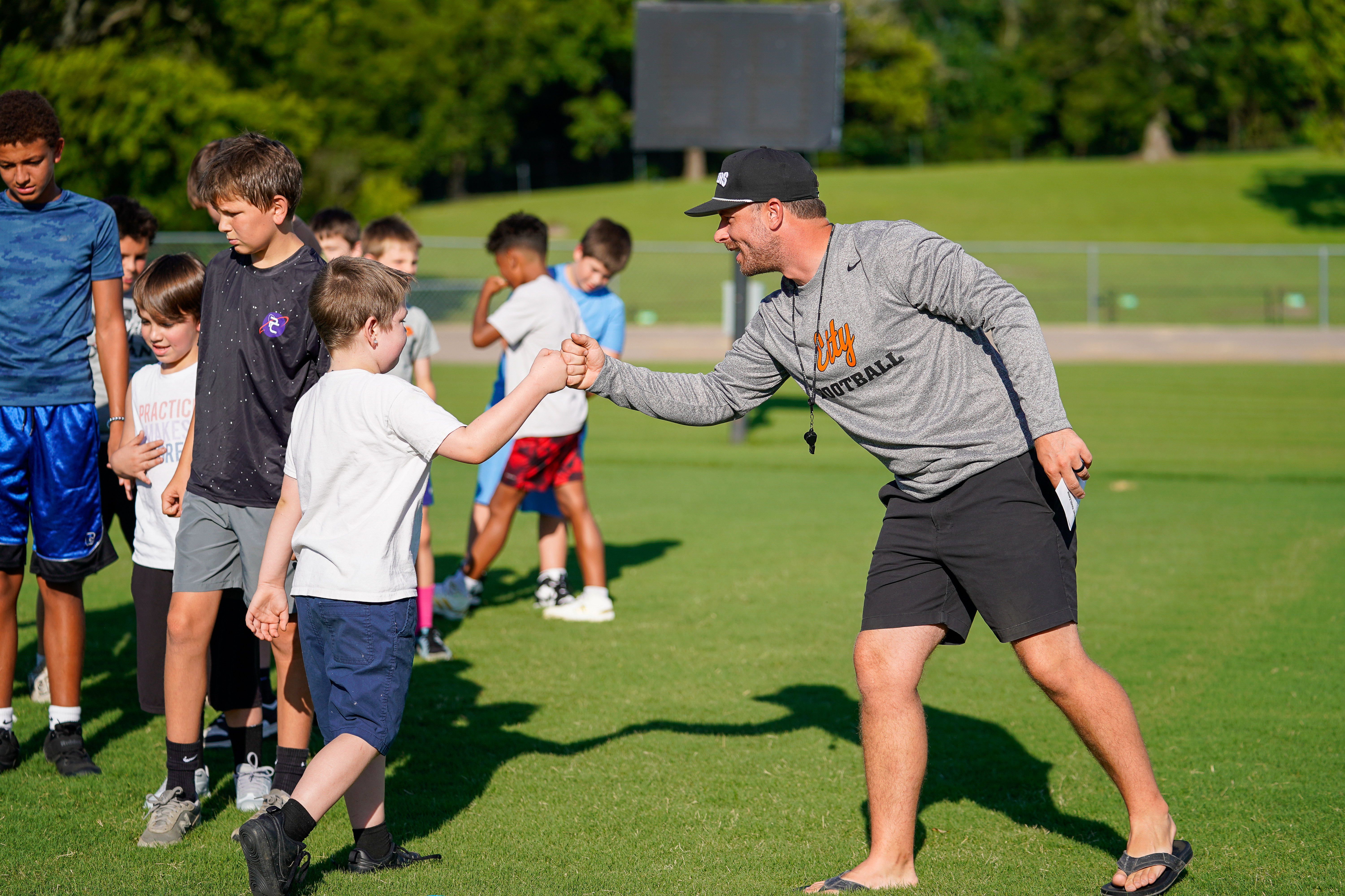 A coach fist bumps a young player on a grassy field with other children in the background and a large screen in the distance.
