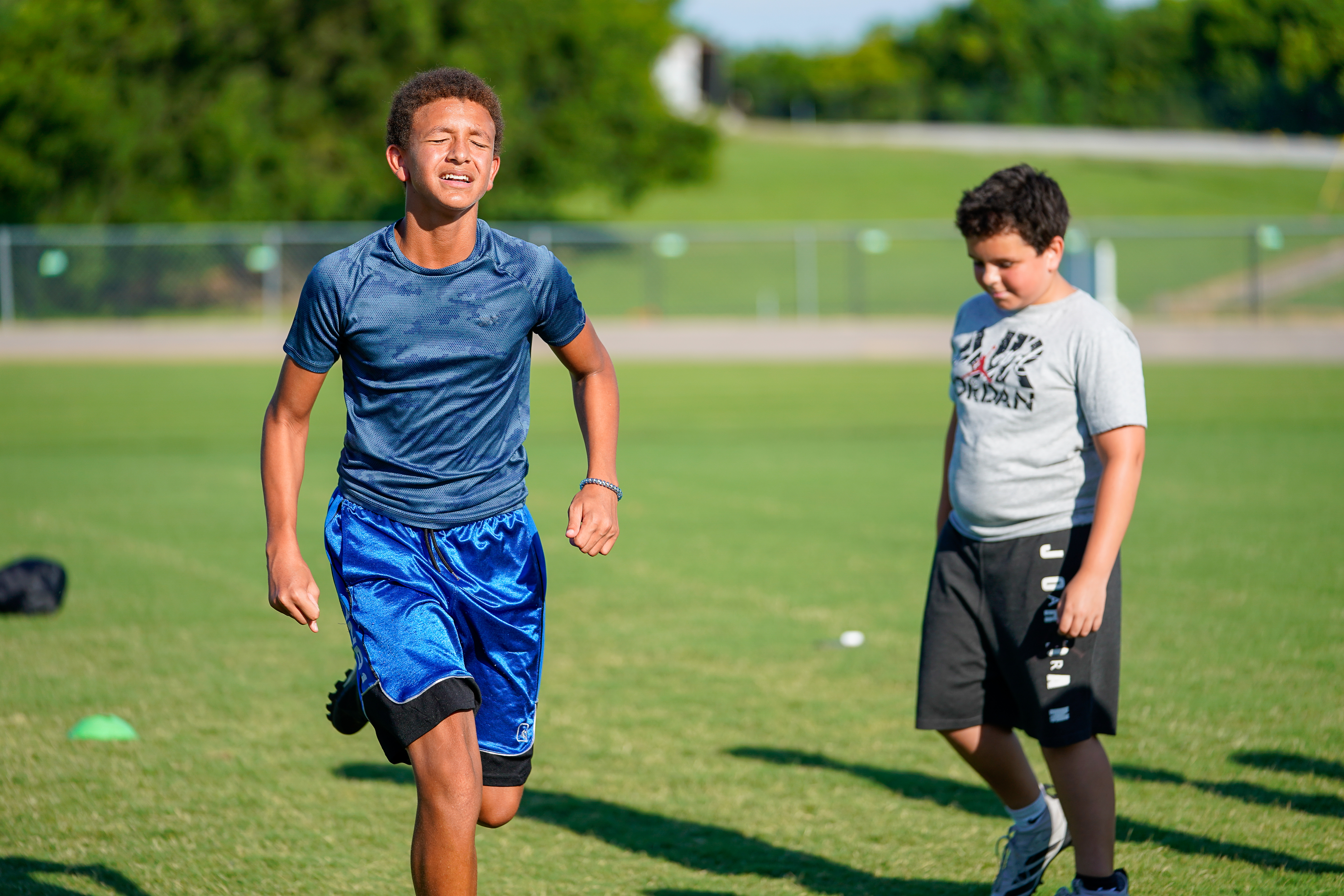 Two young boys are running on a grassy field with trees and a fence in the background.