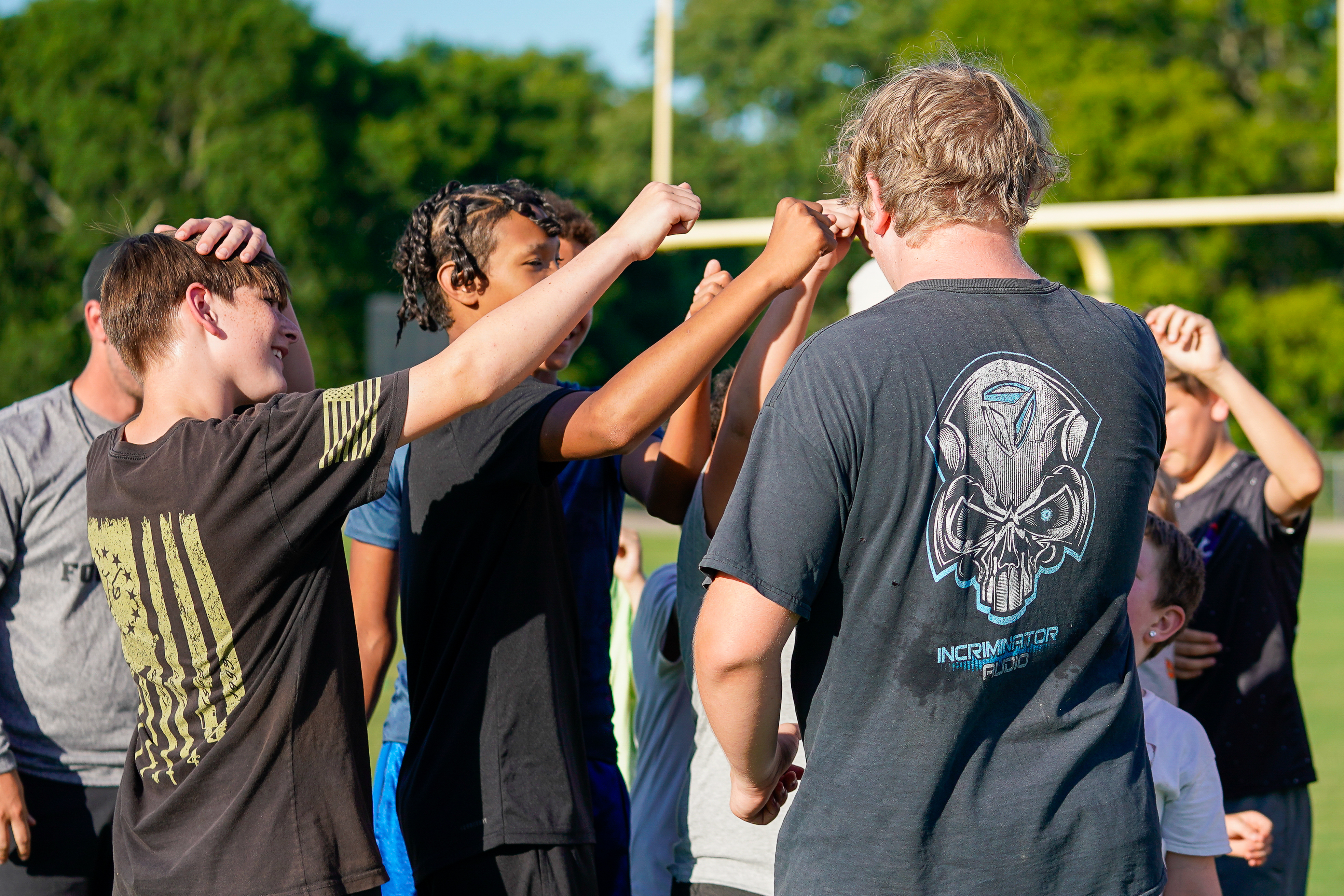 A group of young athletes in the foreground are giving each other fist bumps with a football field and goalposts visible in the background.