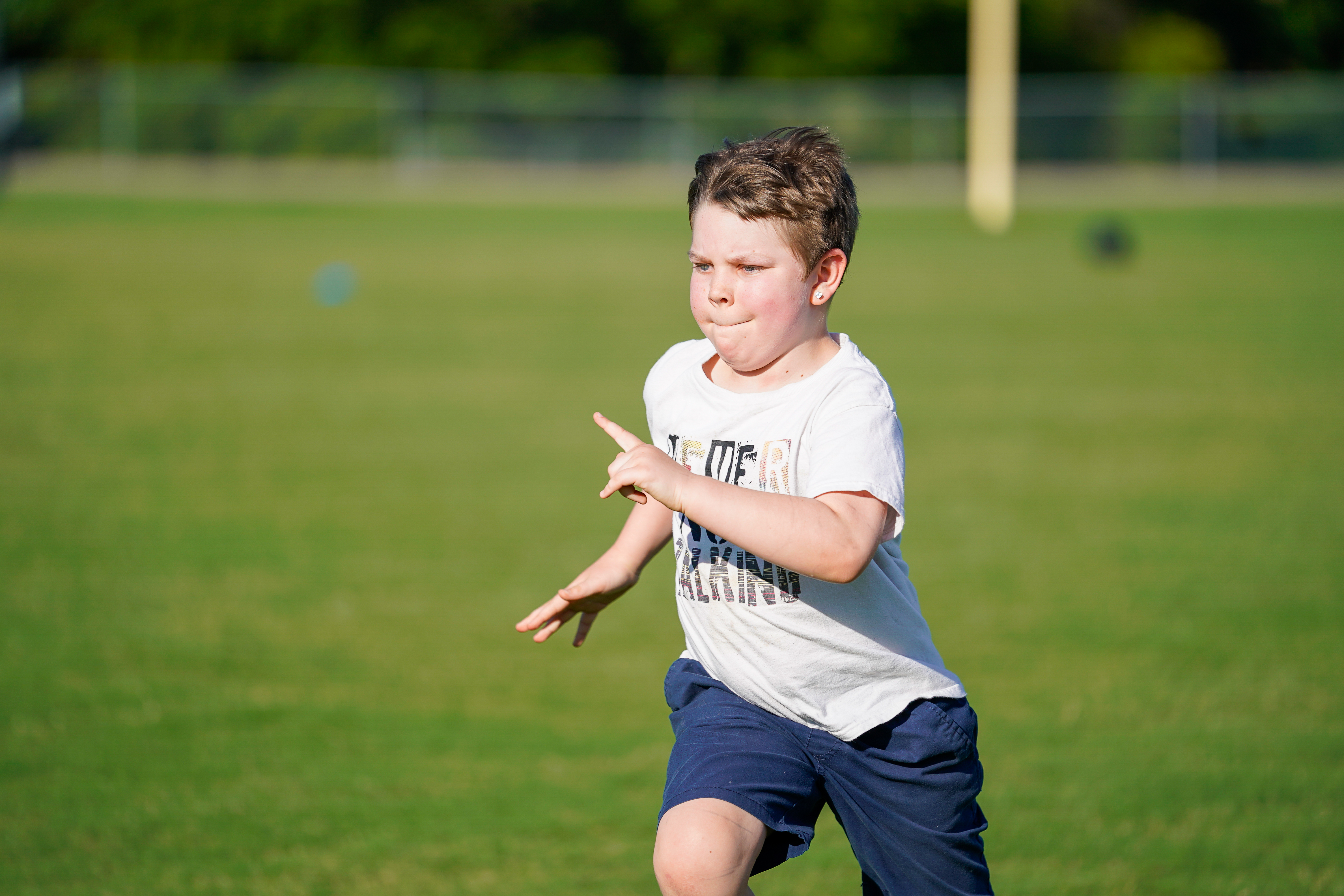 A young boy in a white t-shirt and blue shorts runs across a grassy field with trees in the blurred background.