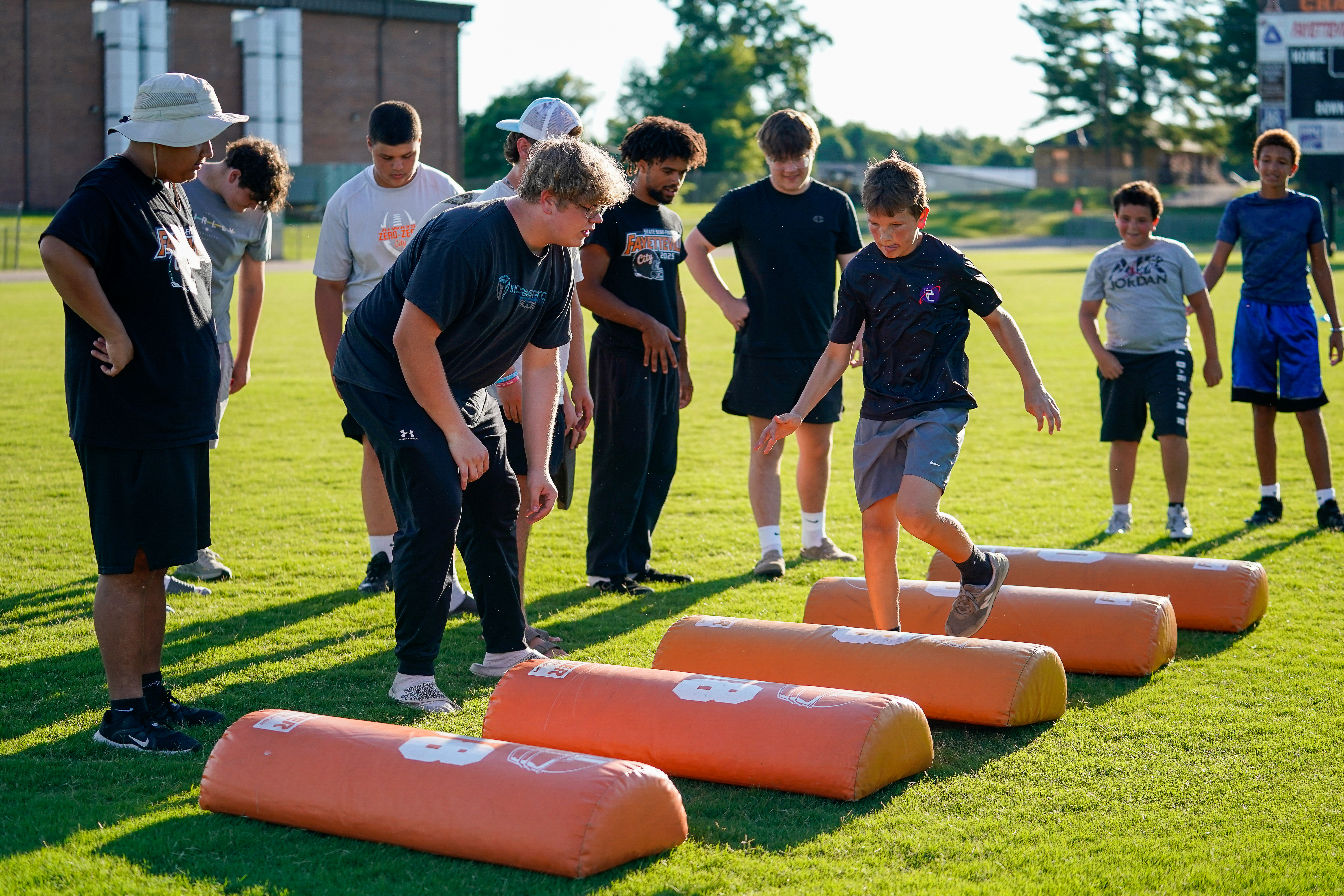 A group of young athletes practice drills on a grassy field with orange blocking dummies in the foreground and a stadium in the background.